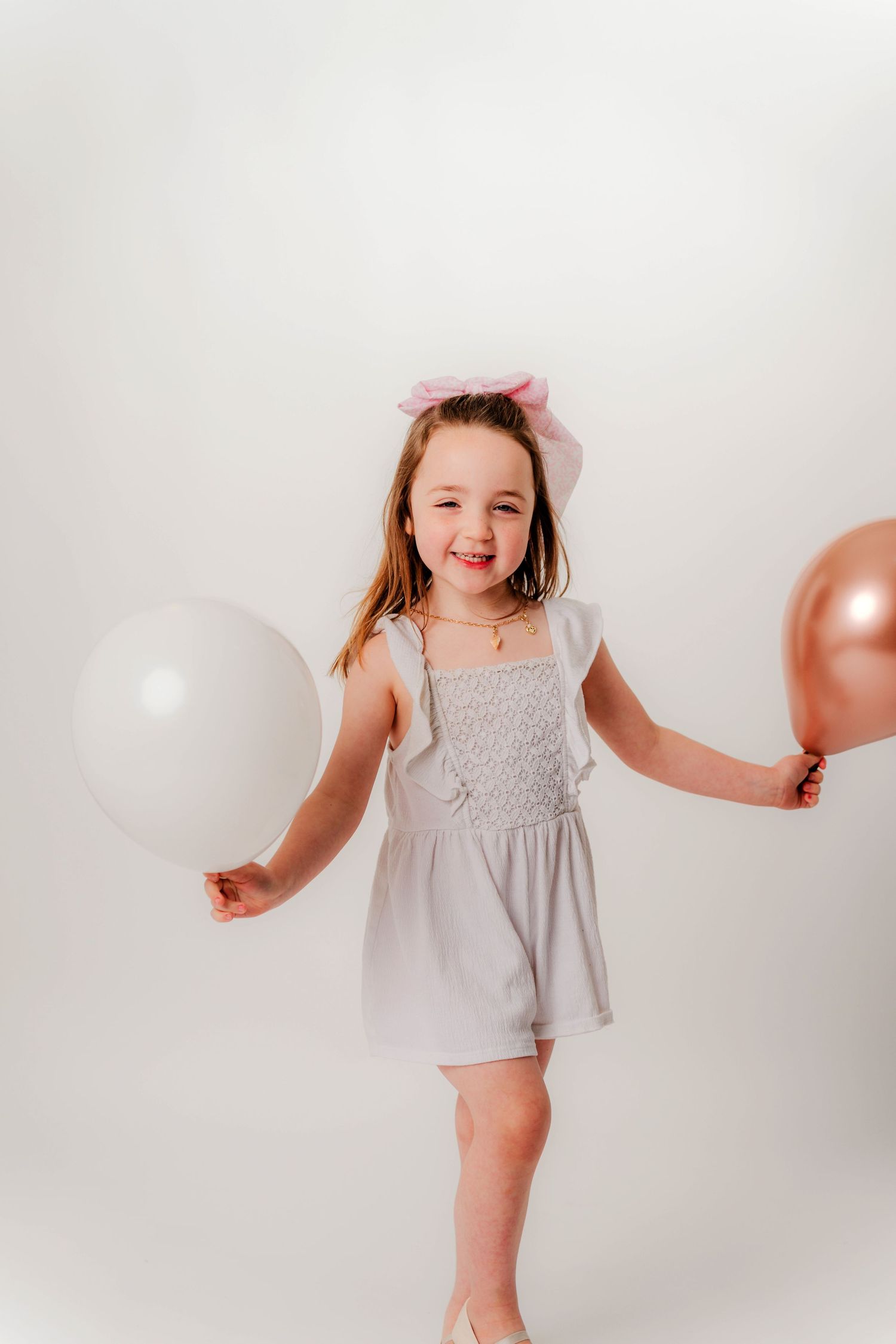 Young figure in light gray dress with pink bow holding white and rose gold balloons against white backdrop.
