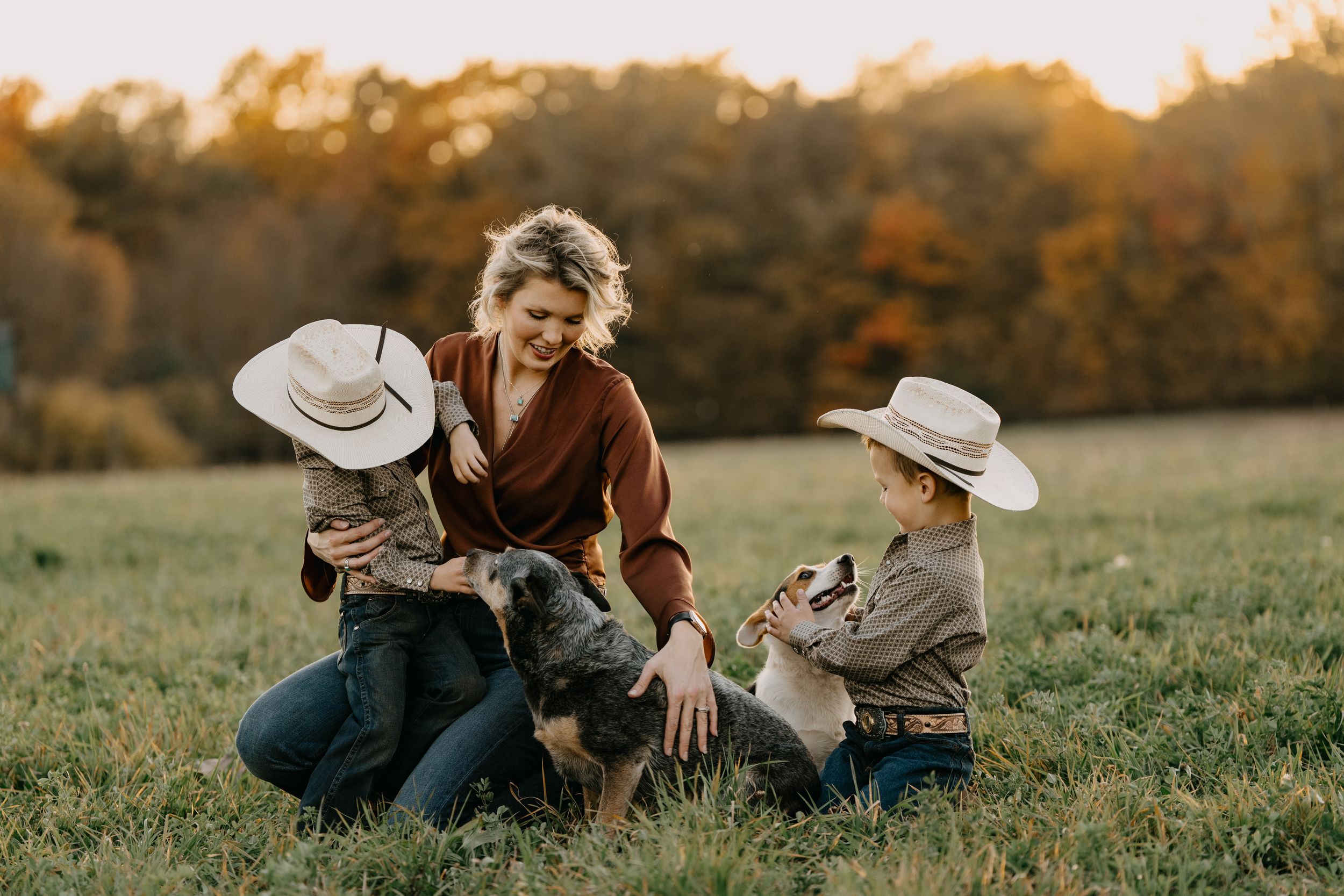 Family in cowboy hats sitting together in a grassy meadow during autumn sunset with their dog.