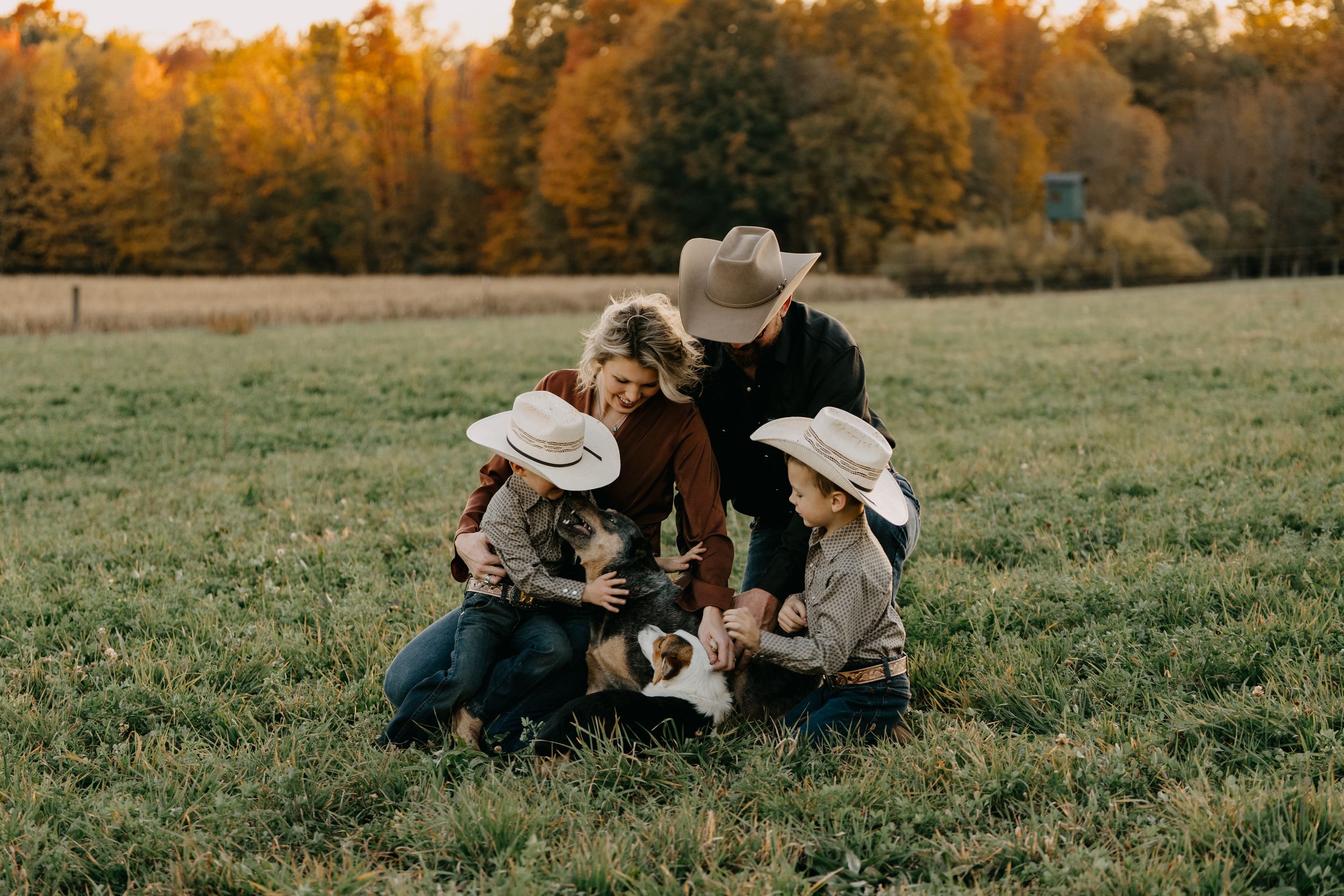A group wearing cowboy hats gathers closely on a grassy field during golden hour with autumn trees in the background.