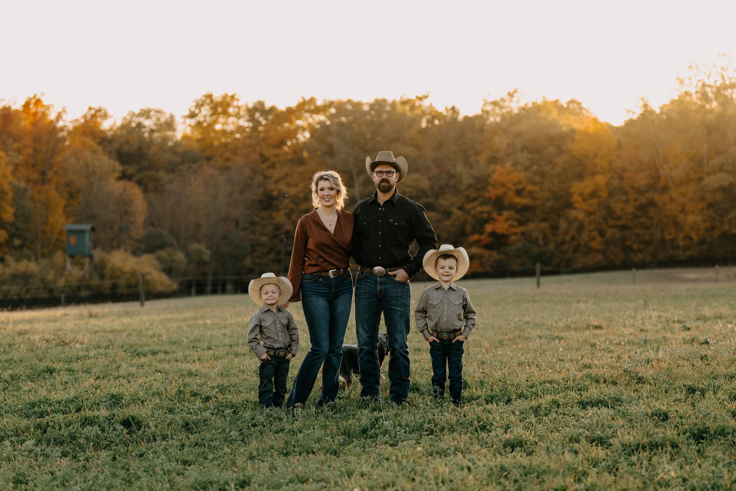 Family of five stands together in an autumn field at sunset with golden light filtering through trees in background.