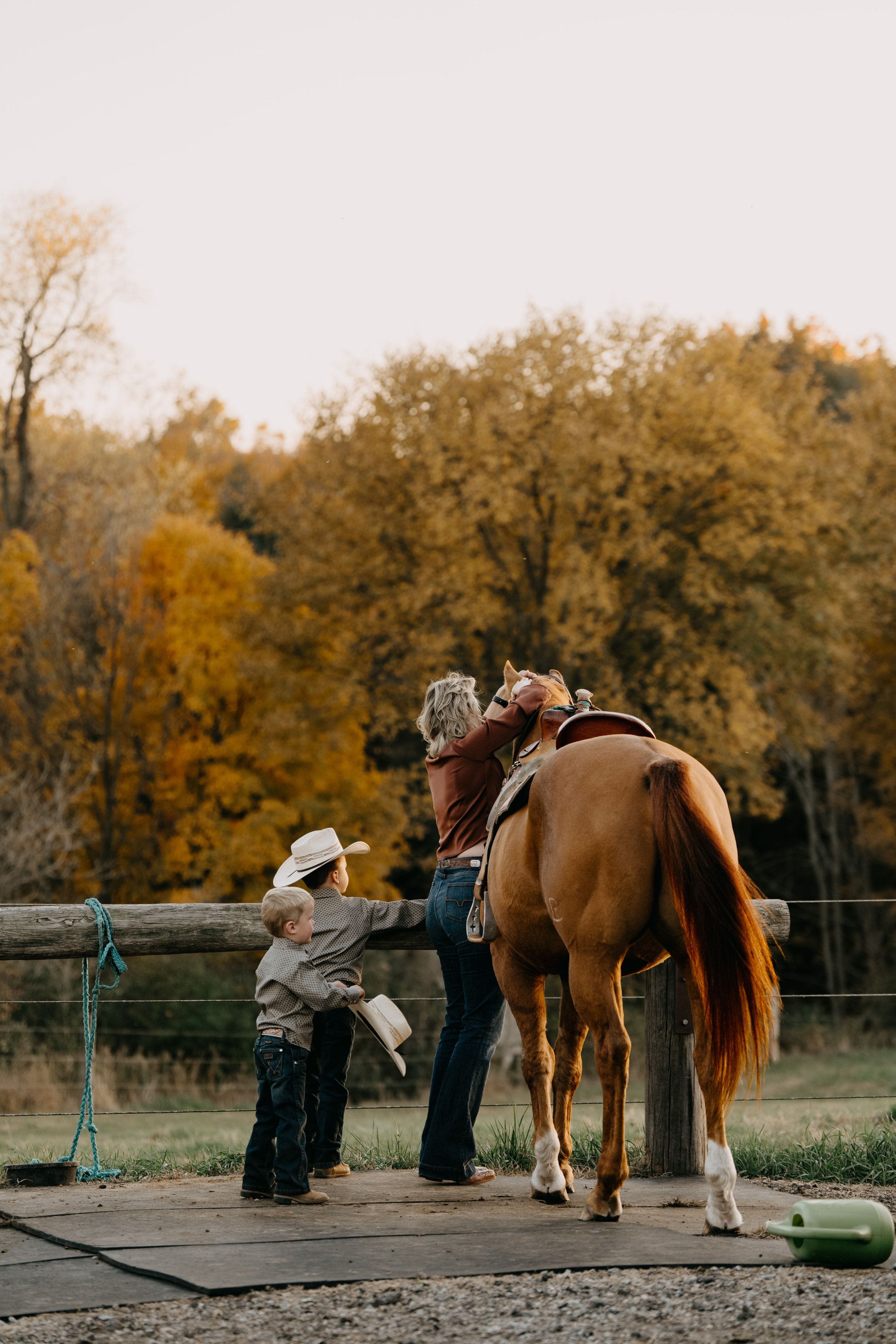 A chestnut horse stands on a platform at a ranch while people prepare for a horseback riding lesson on an autumn day.