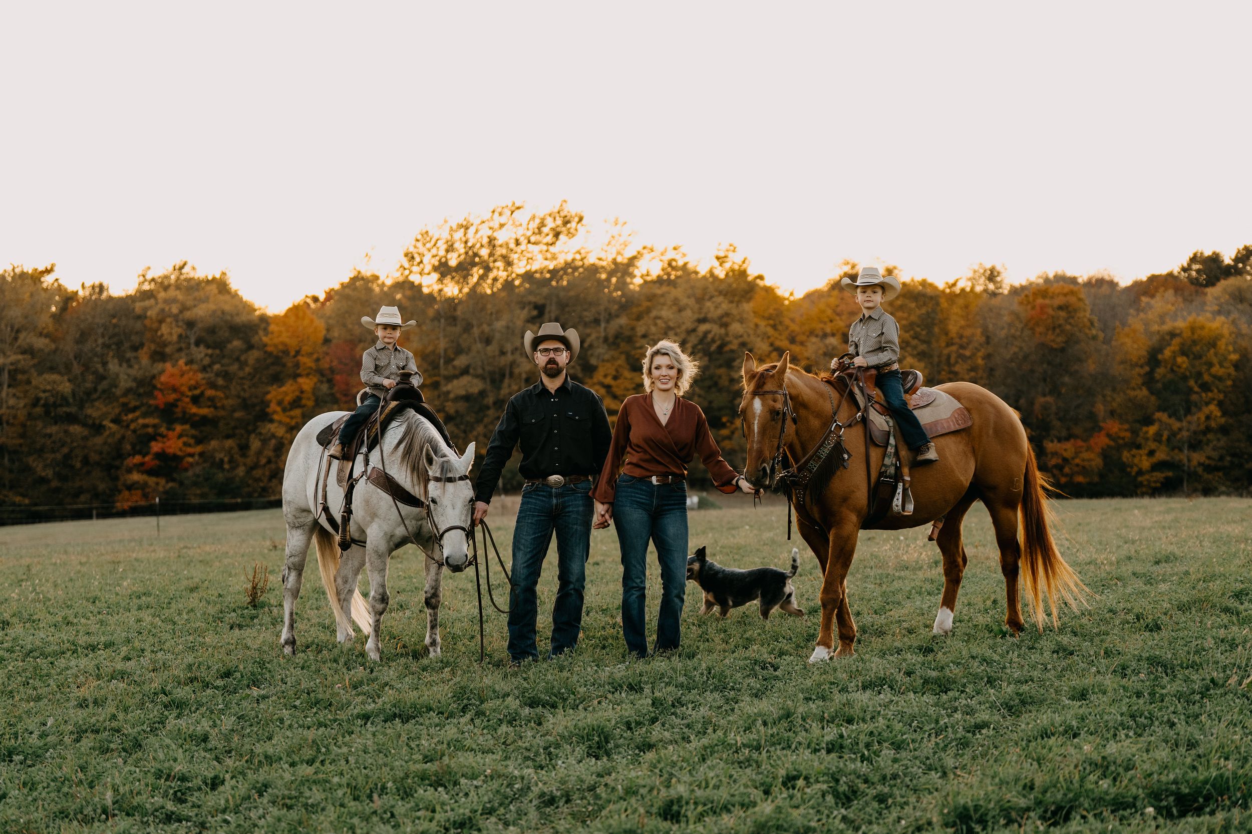 Two horses and three people standing together in a grassy field during golden hour with autumn trees in background.