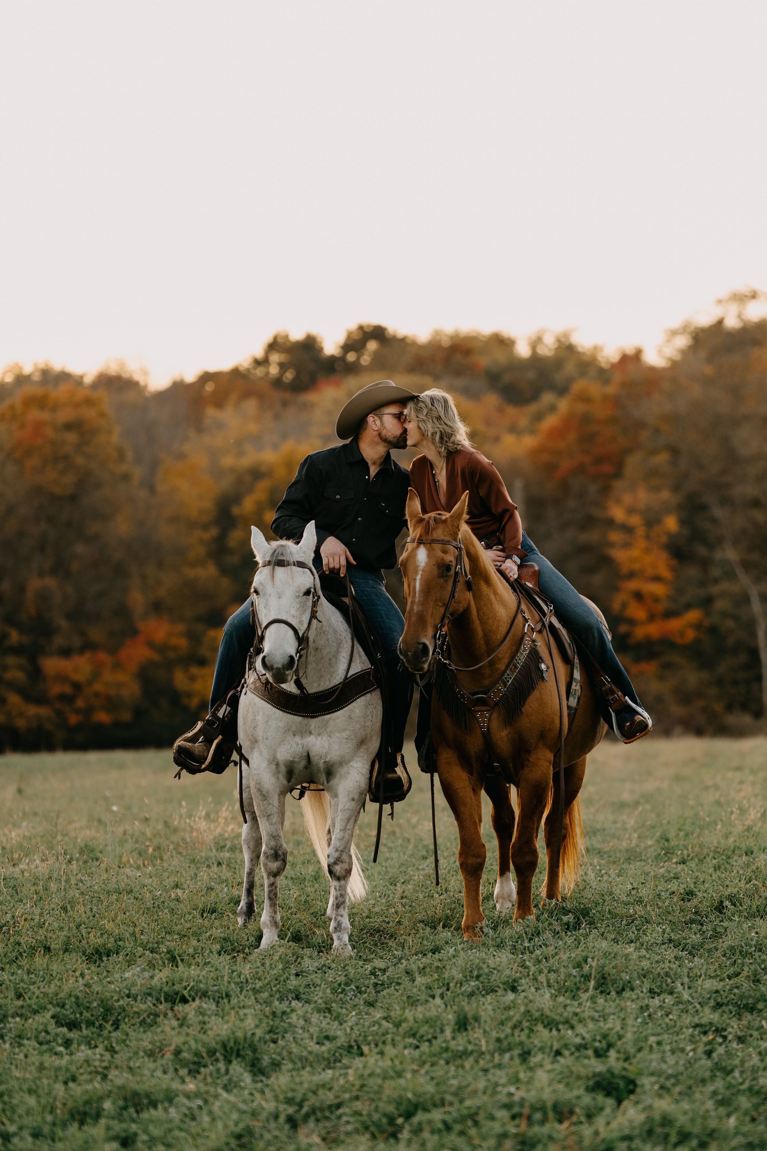 Two equestrians riding horses side by side on grassy meadow with autumn foliage in the background at sunset.
