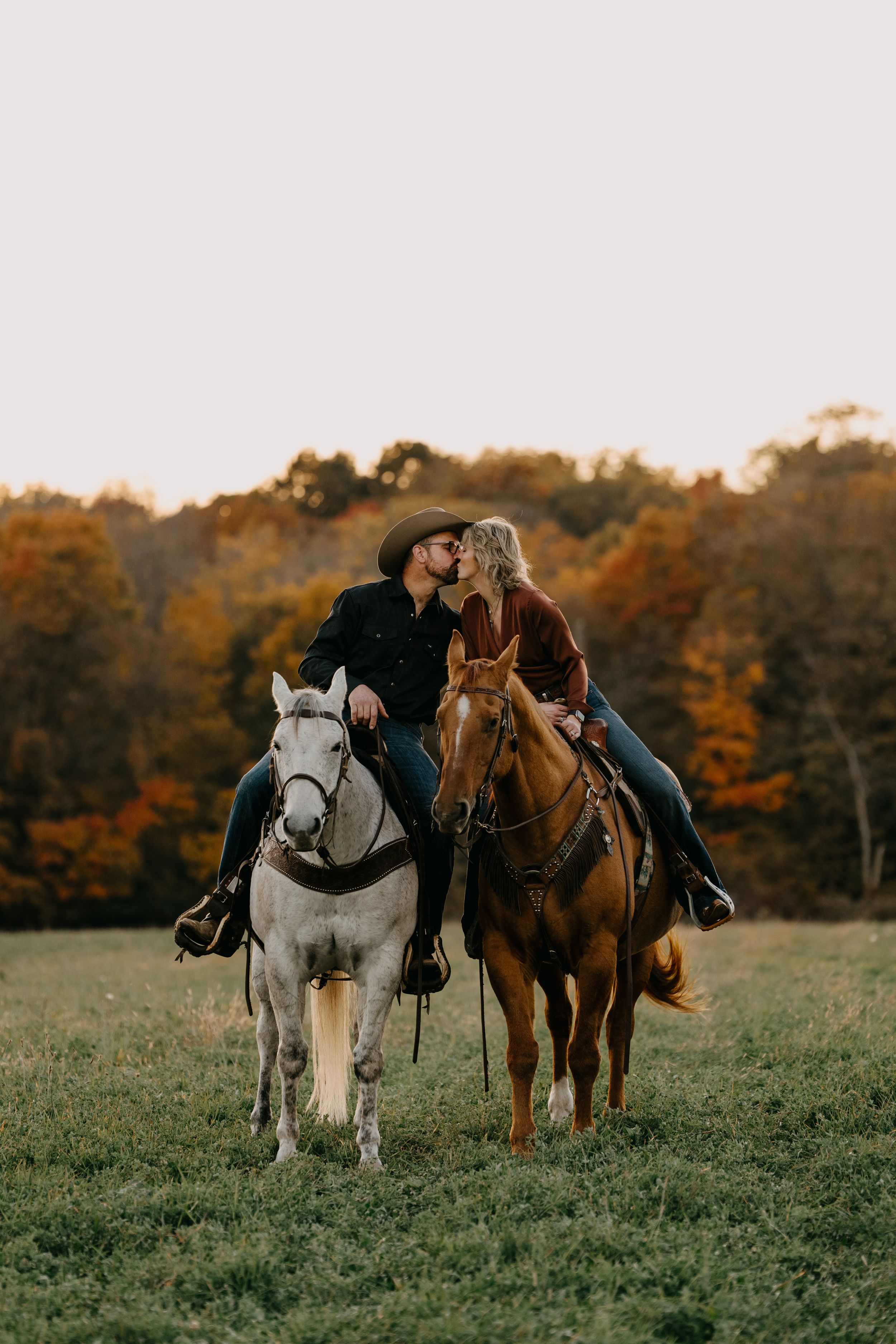 Two riders on horseback share a moment together in an autumn field with orange and yellow foliage in the background.