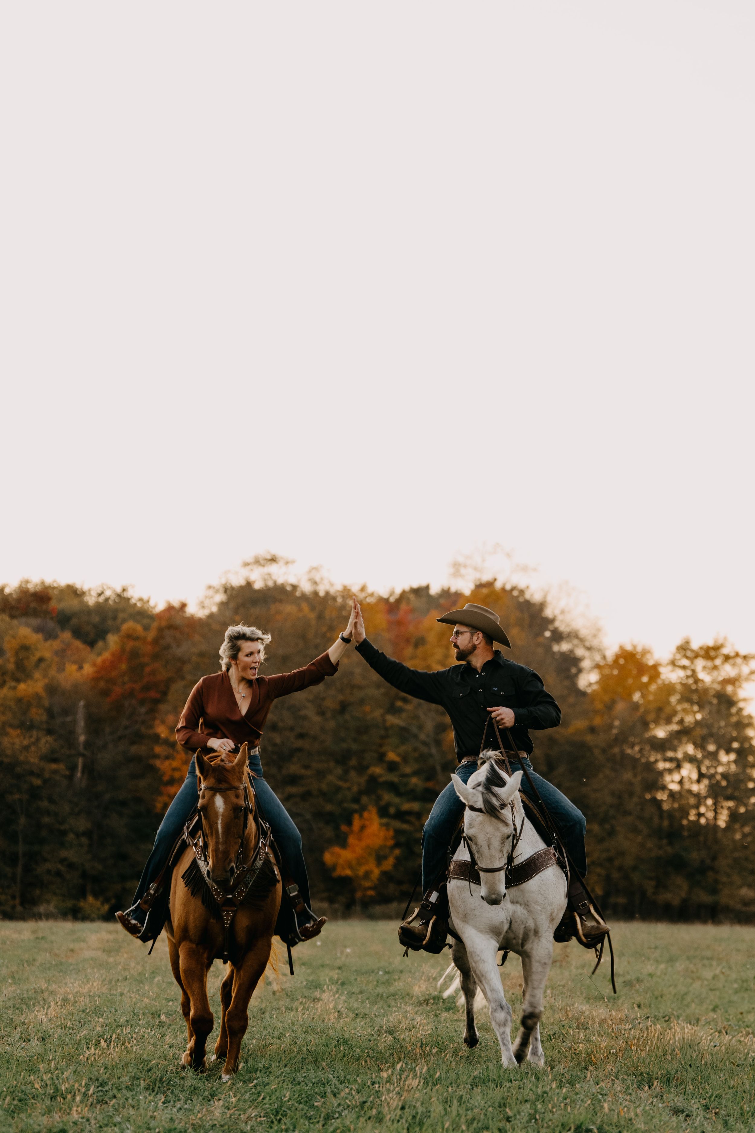Two people riding horses side by side giving each other a high five in an autumn field at sunset.
