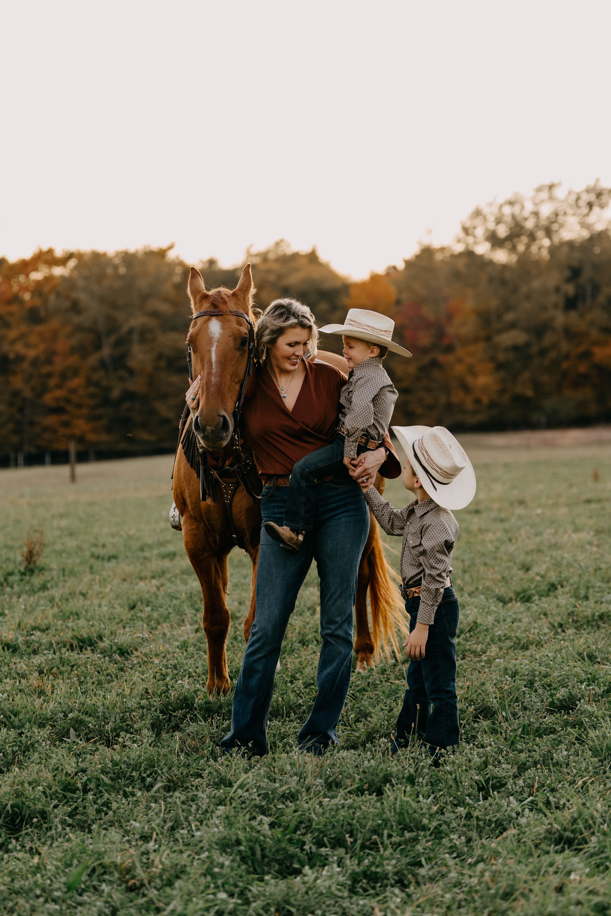 Two people in western attire stand with a chestnut horse in an autumn field during golden hour sunset.