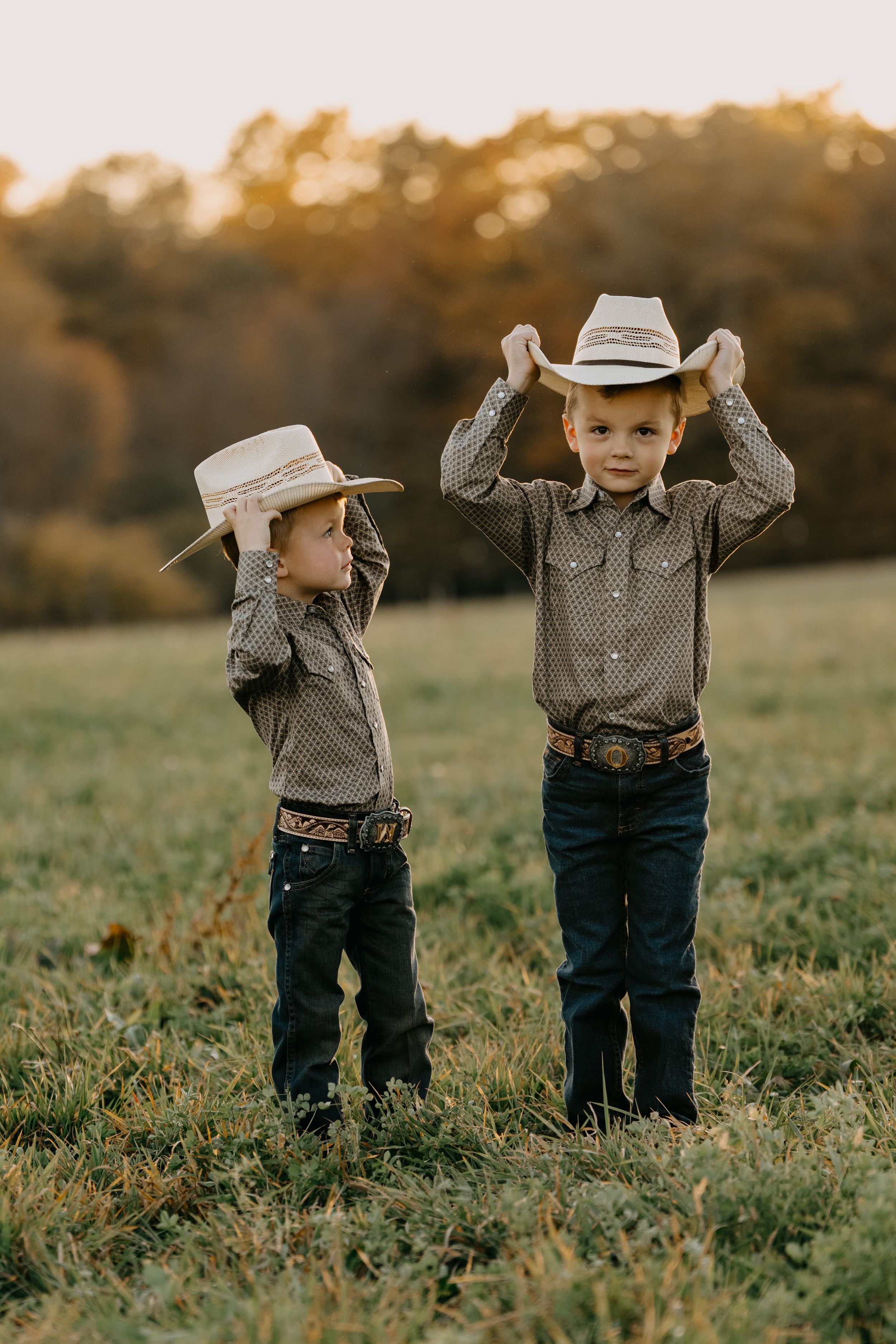 Two young children in western wear and cowboy hats pose together in a grassy field at sunset.