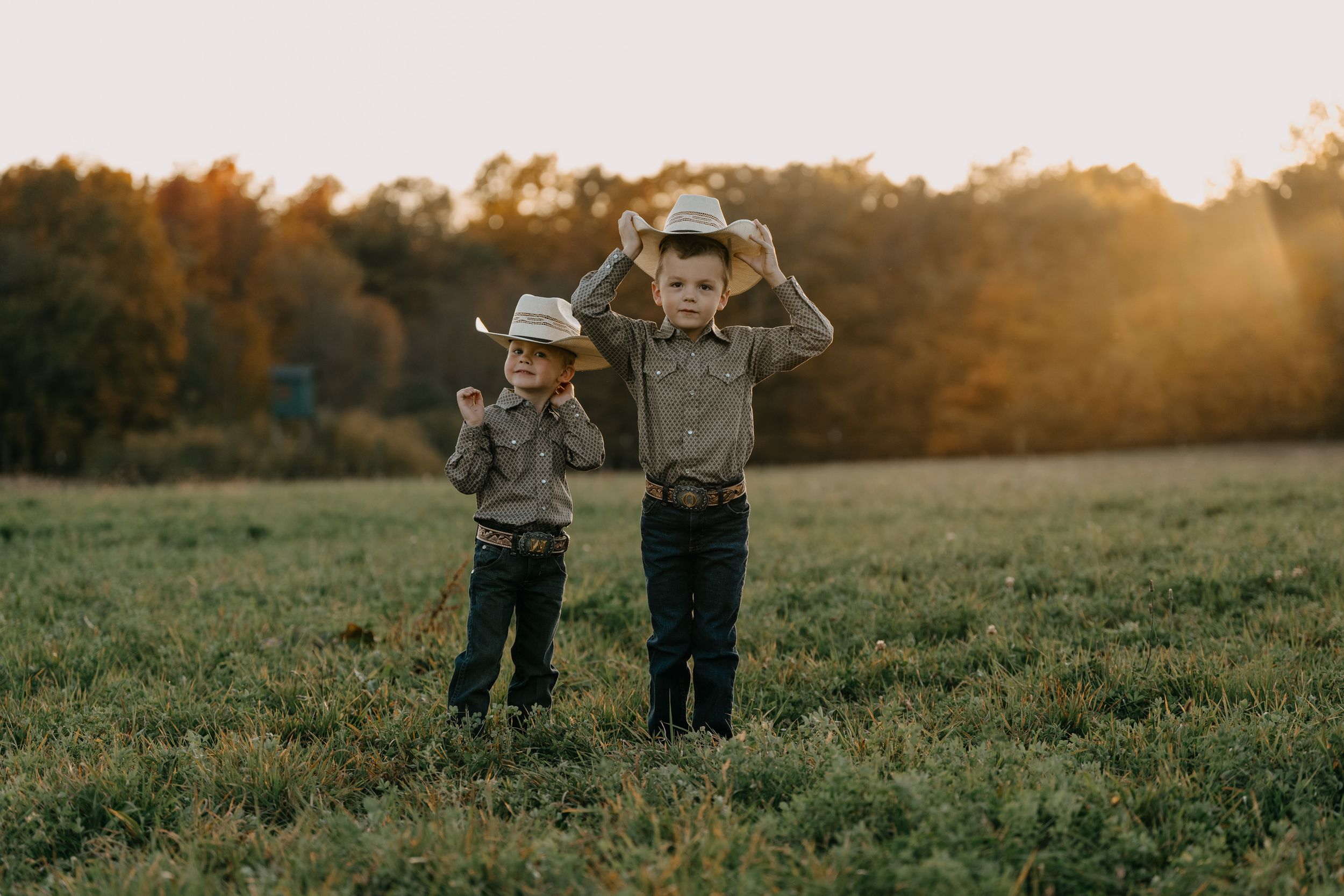 Two children wearing cowboy hats standing together in a field at golden sunset.