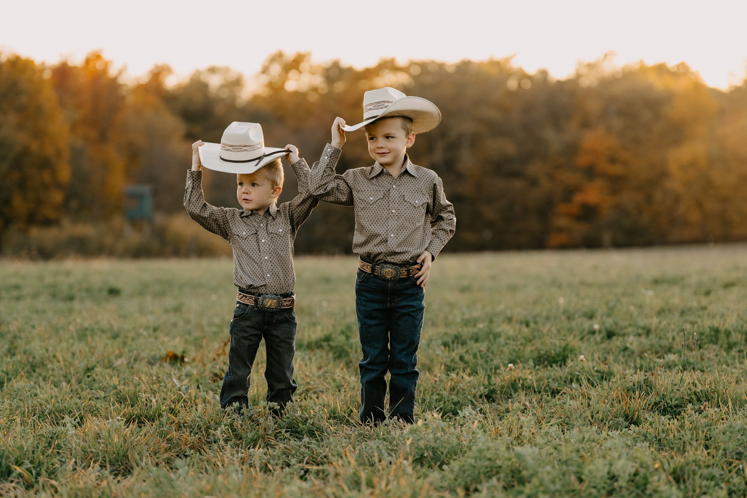 Two young cowboys in western attire stand together in a grassy field at sunset during golden hour.