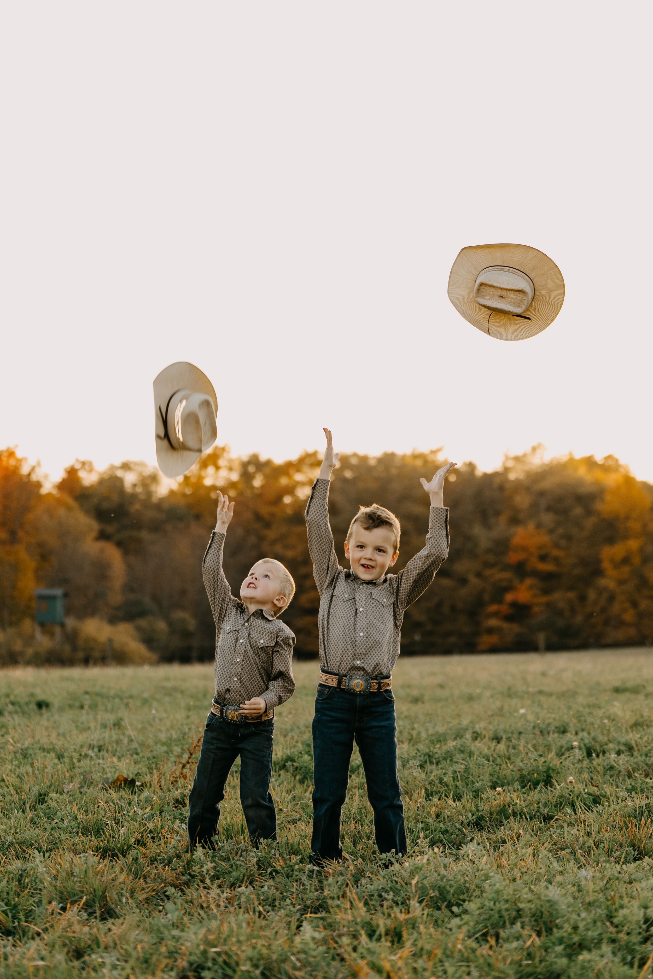 Two people in rustic clothing toss straw hats into a golden autumn field at sunset.