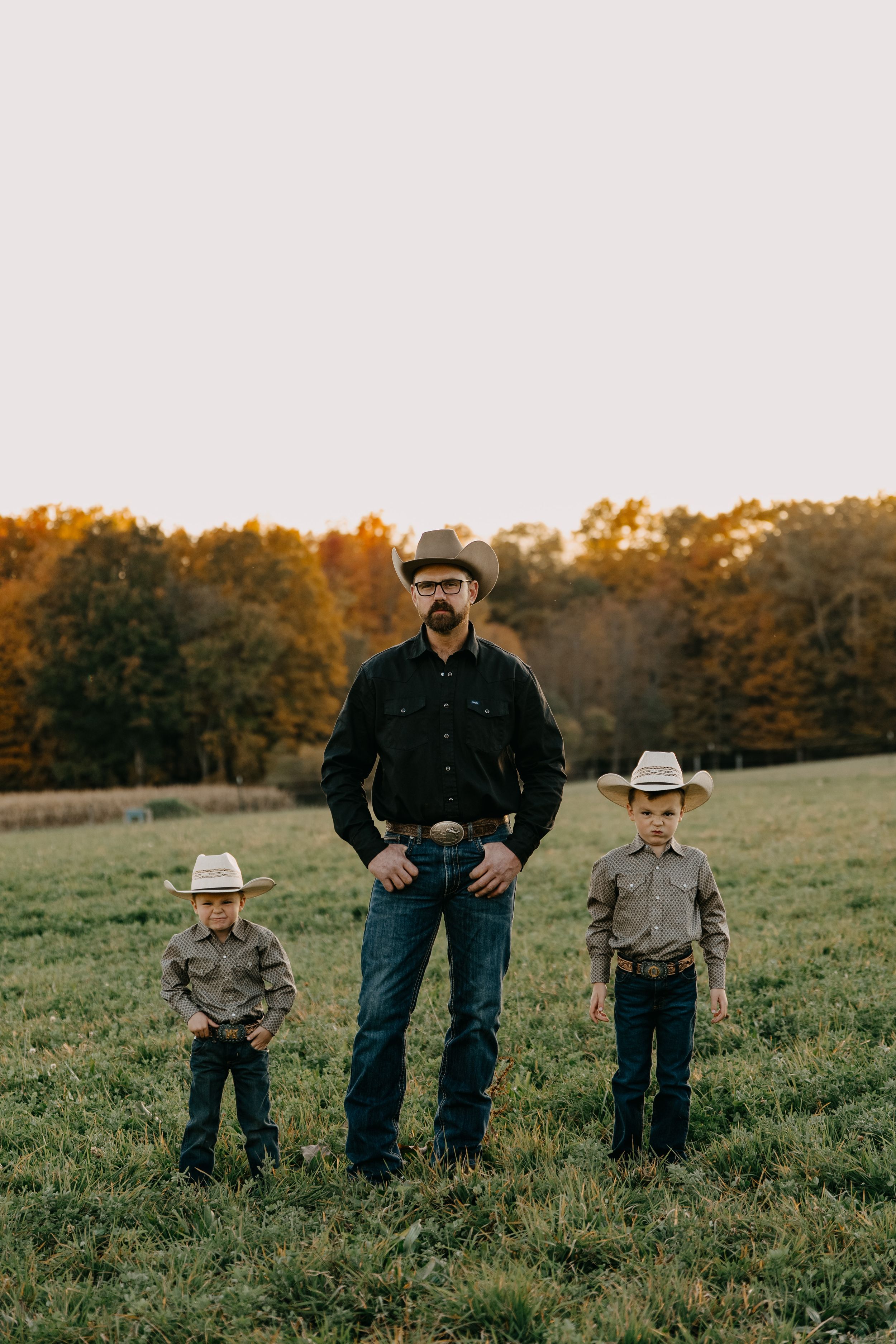 Three people wearing cowboy hats stand together in a grassy field during golden hour with autumn trees in background.