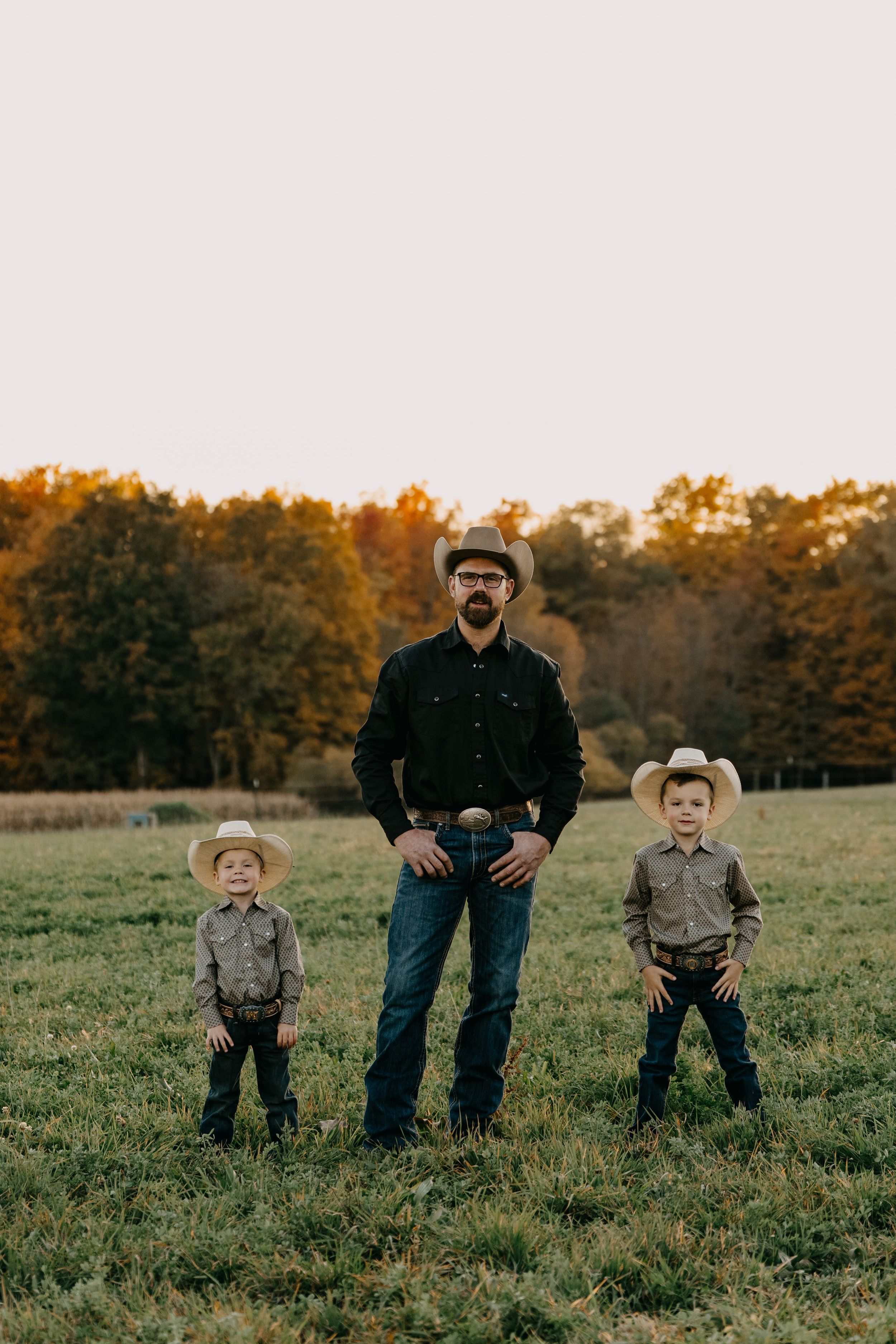 A western-styled family portrait in an autumn field with golden trees in the background during sunset.