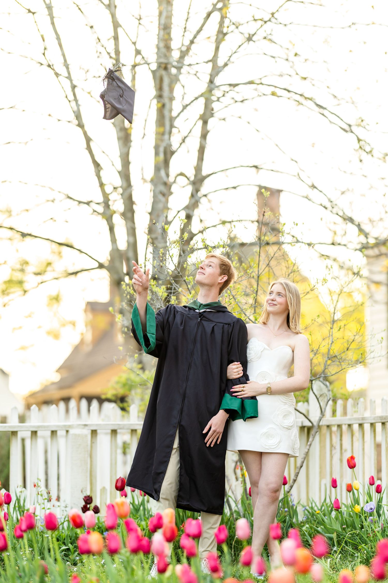 Light and airy photography of William and Mary graduate throwing cap in air. Posed with girlfriend on his arm in a tulip garden.