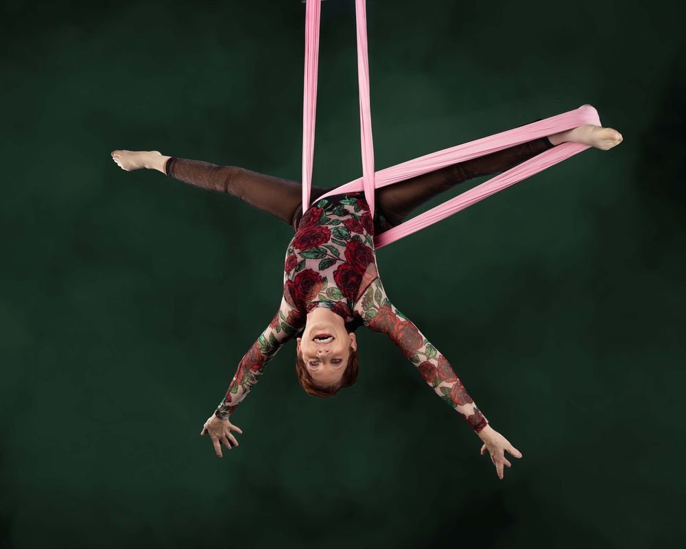 An aerial acrobat performs graceful suspended movements in a dimly lit space using pink fabric ribbons.