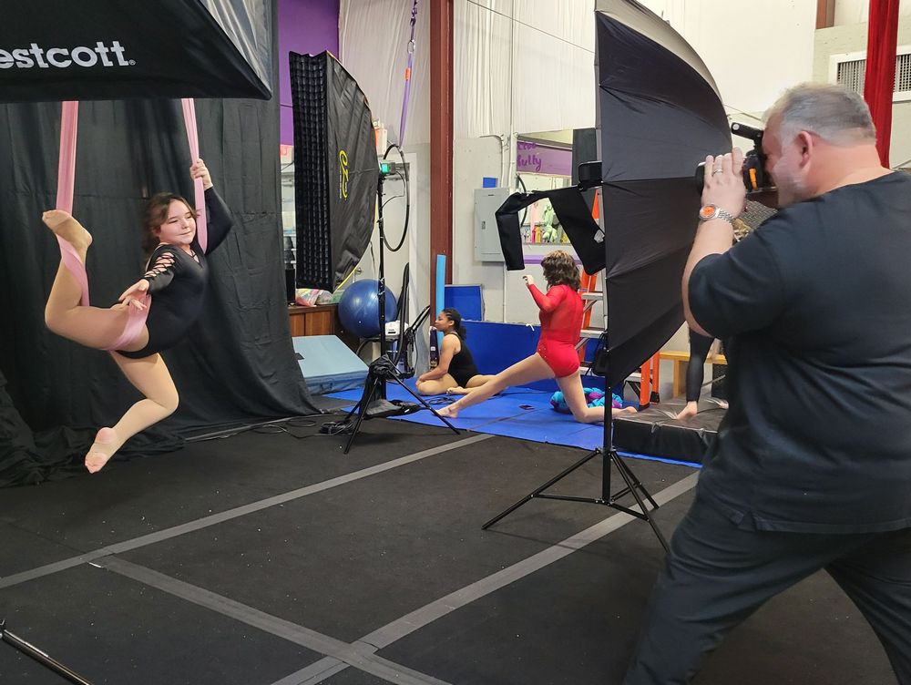 A photographer captures an acrobatic performer suspended from straps during a photo shoot at a fitness studio.