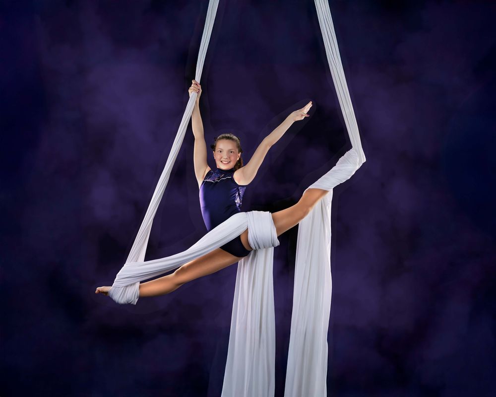 An aerial acrobat performs graceful poses on white silk ribbons against a dark backdrop in a performance sequence.