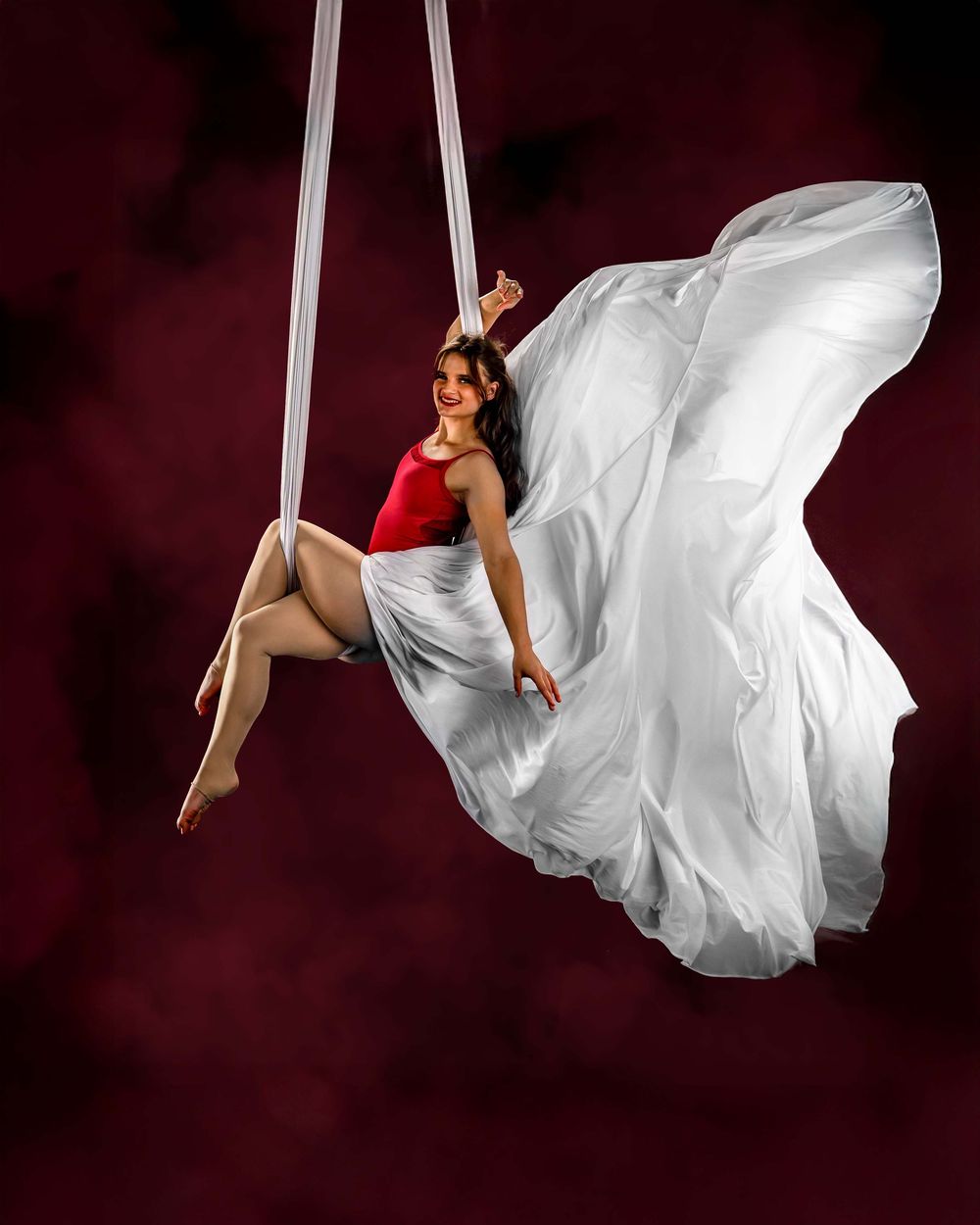 An aerial performer in a red top and flowing white fabric dress gracefully suspended on silk ribbons against a dark background.