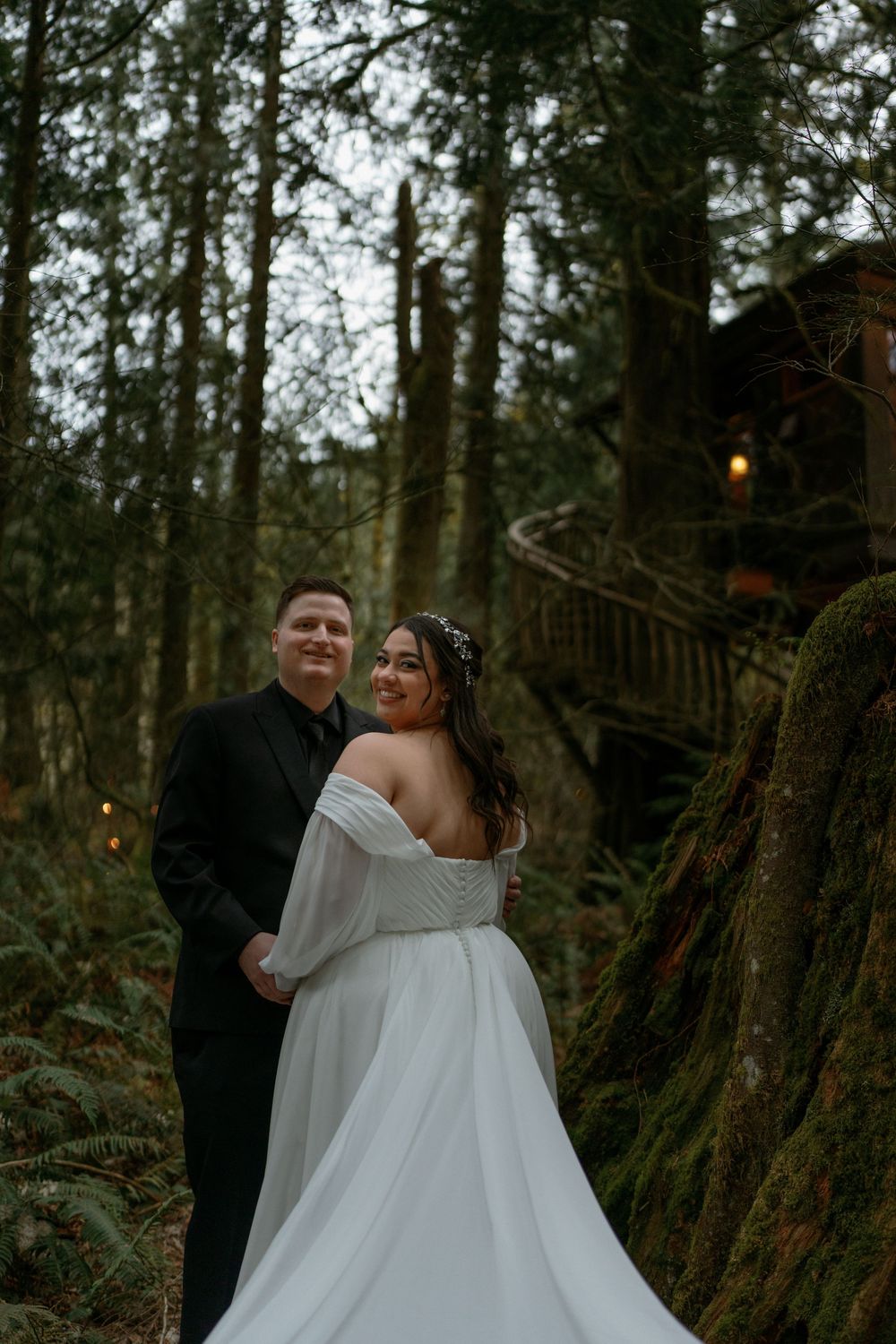 A couple shares a romantic moment in a misty forest with wooden stairs and twinkling lights in the background.