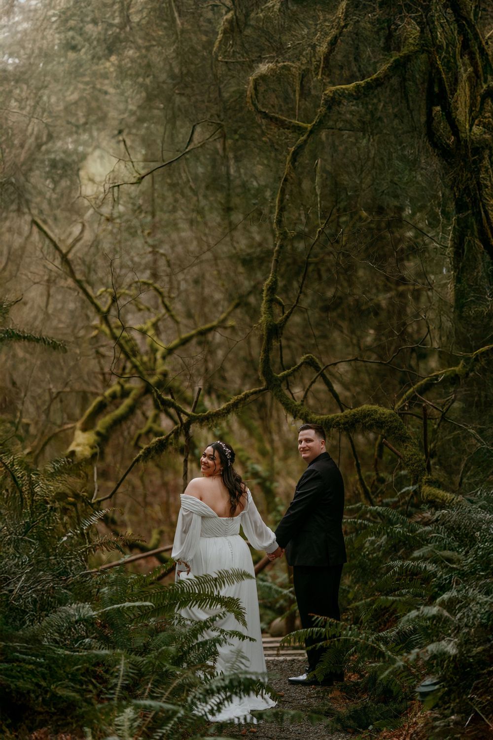 A couple in wedding attire share a joyful moment while walking through a misty forest path surrounded by ferns.