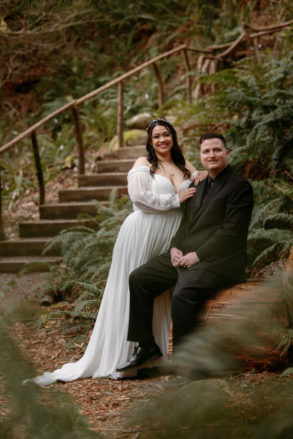 A couple in formal wedding attire sits together on rustic stone steps surrounded by lush greenery in a forest setting.
