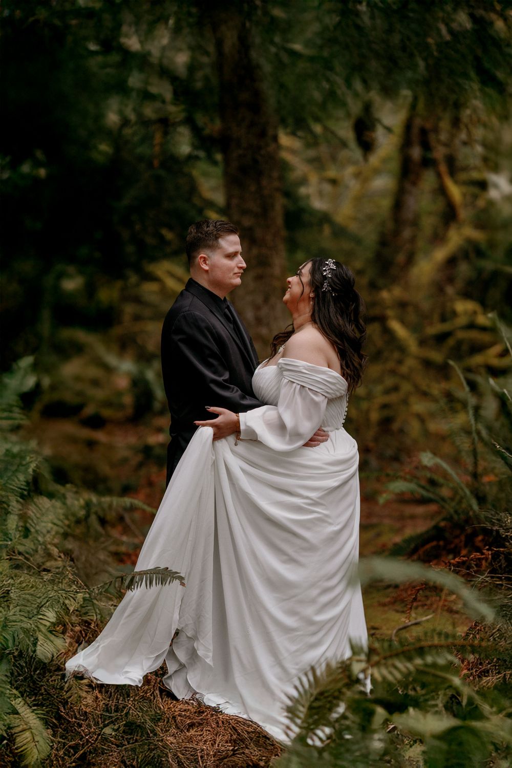A romantic woodland wedding photo shows a couple embracing among ferns and trees in moody forest lighting.