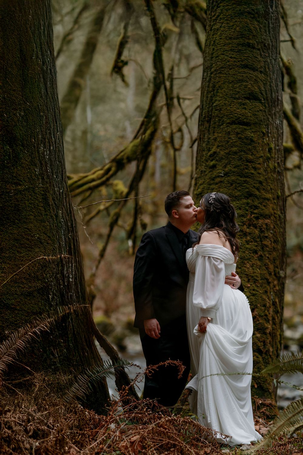A couple shares an intimate moment in a misty forest setting, surrounded by moss-covered trees and fallen leaves.