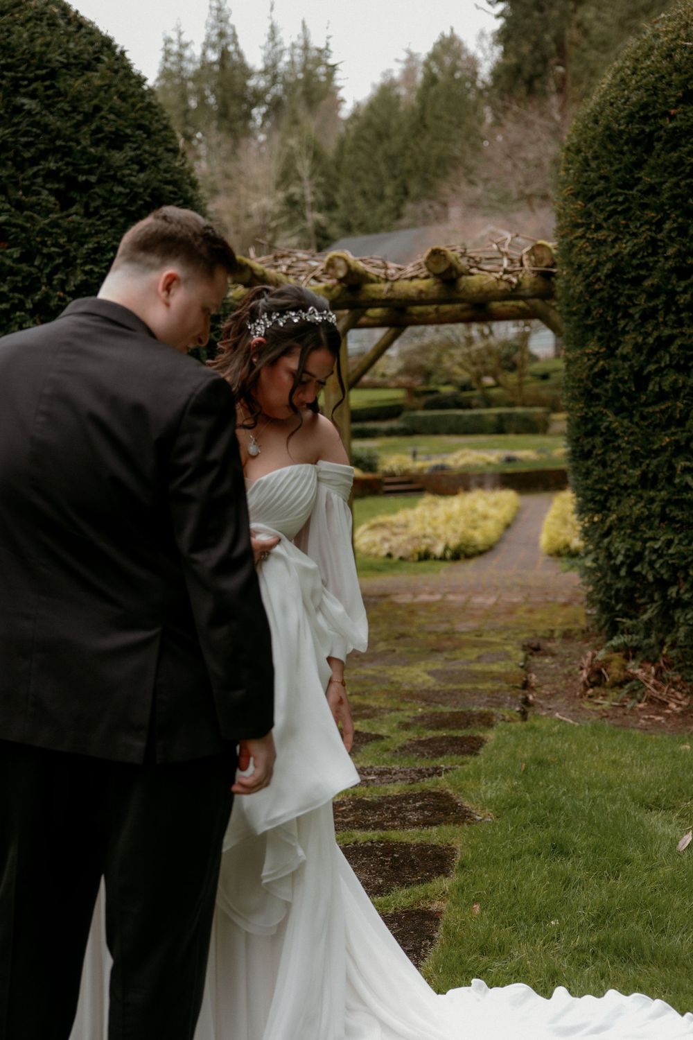 A couple in wedding attire share an intimate moment in a lush garden with manicured hedges and stone pathways.