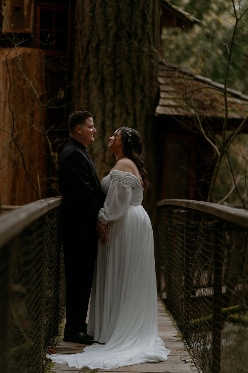 A romantic wedding couple embraces on a rustic wooden bridge surrounded by forest trees in moody lighting.