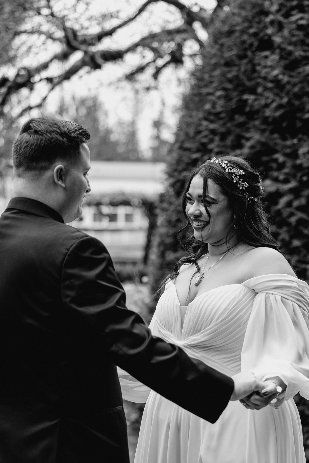 Black and white photo of a wedding couple sharing a joyful moment in a garden setting with lush hedges in the background.