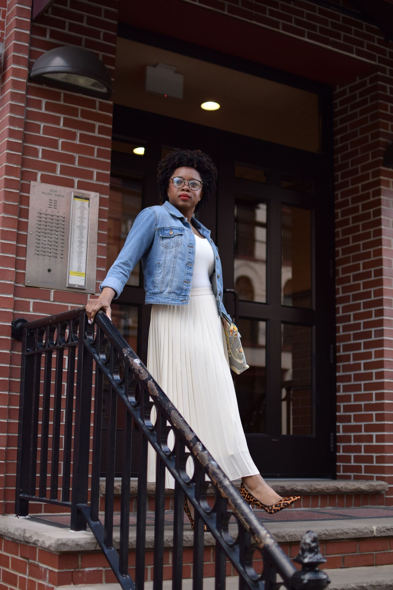 Person in light colored clothing and denim jacket stands at entrance of brick building with black metal railing.