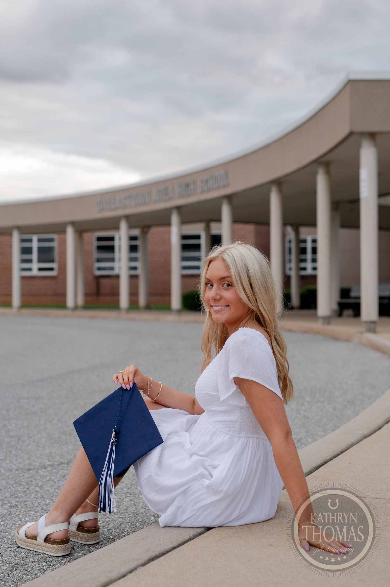 Graduate in white dress sitting on curb with diploma at curved building entrance with columns on cloudy day.