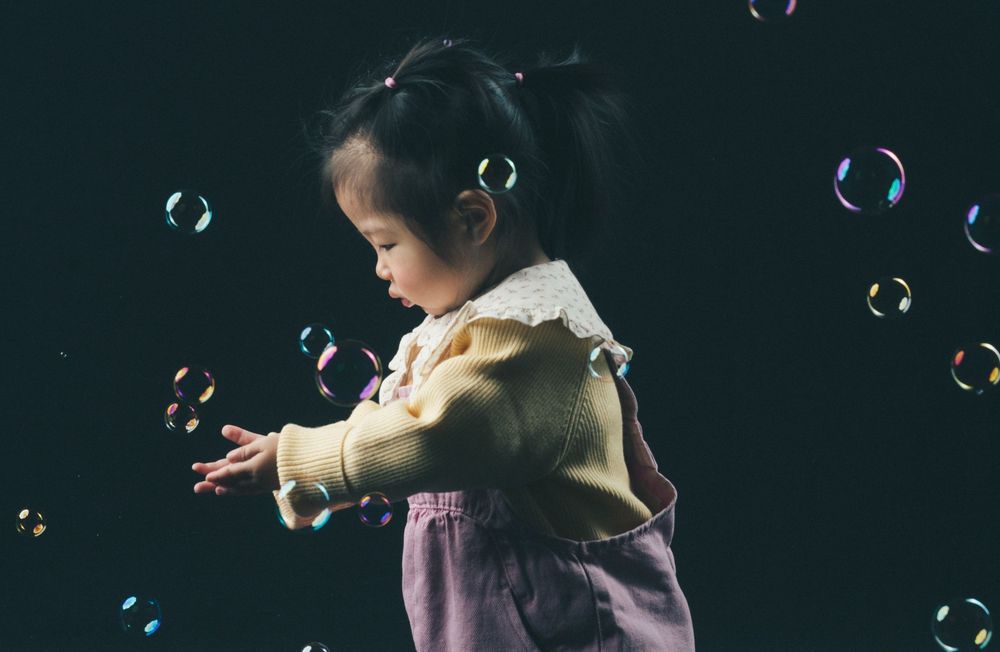 Child in traditional dress playing with soap bubbles against dark background.