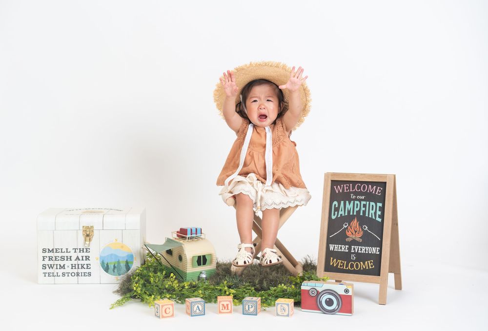 Playful child sits surrounded by camping-themed props and decorative elements in photography setup.