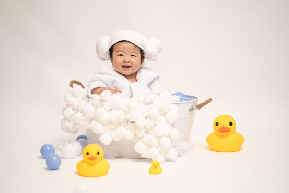 Baby in white outfit sits in bubble bath setup with yellow rubber ducks for studio photography.