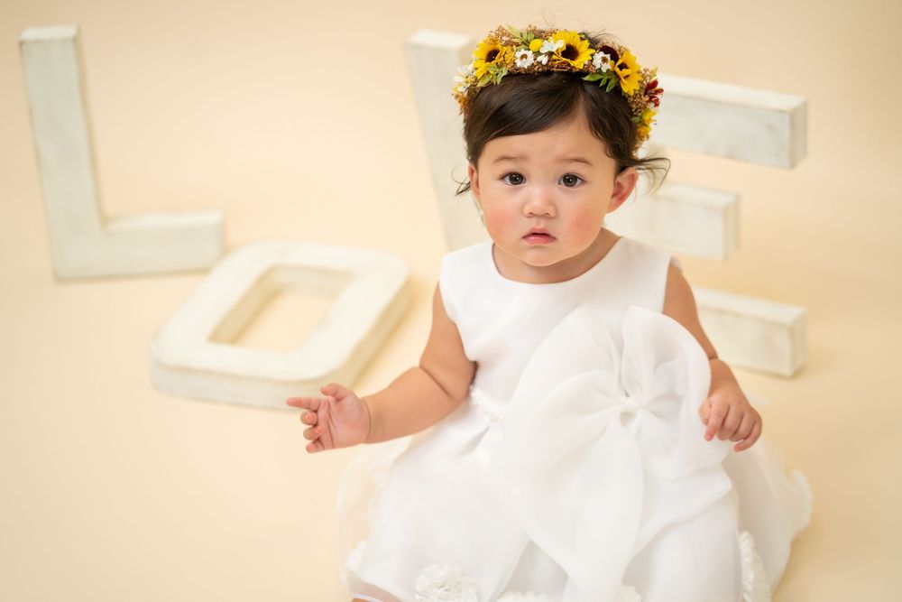 Child in white dress with yellow flower crown against light background.
