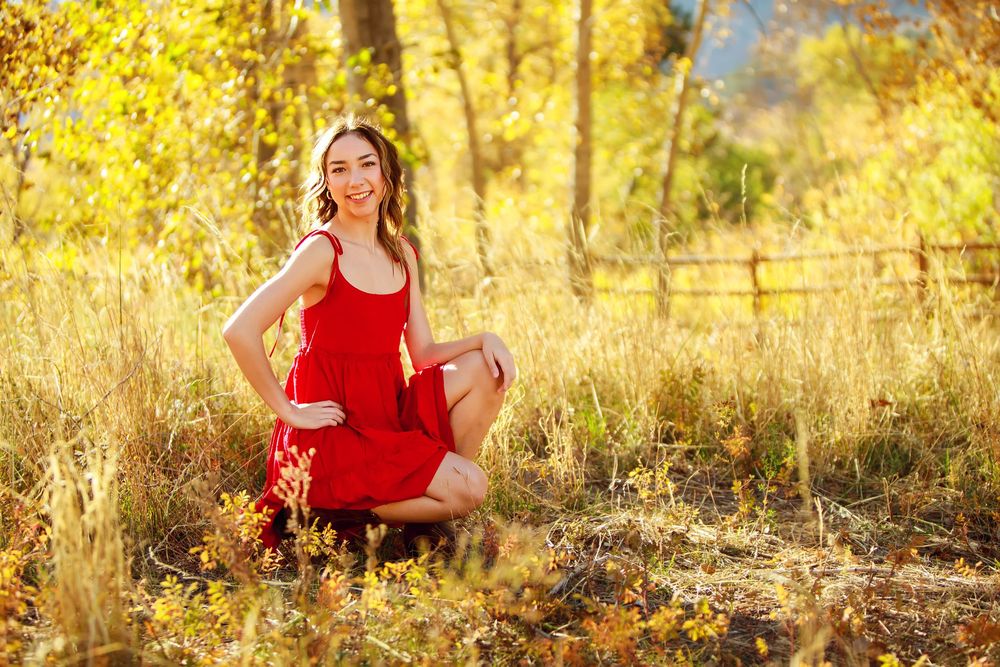 Woman in red dress poses gracefully in golden autumn field with fence in background.
