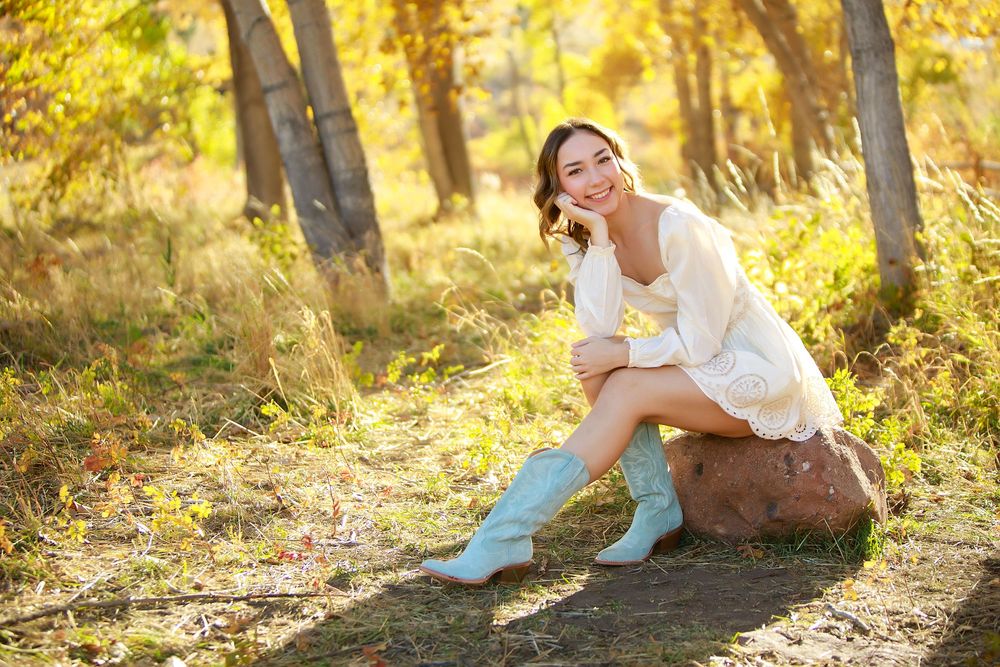 Person in white dress and blue boots sitting on rock in sunlit autumn forest.