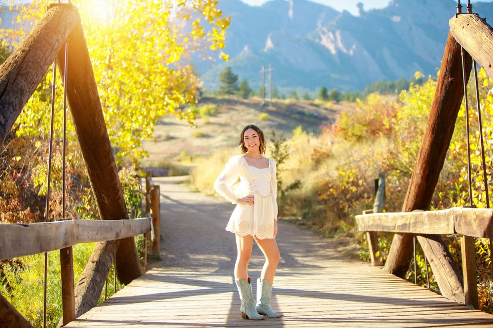 Person in white dress posing on wooden bridge with mountain backdrop and autumn foliage.