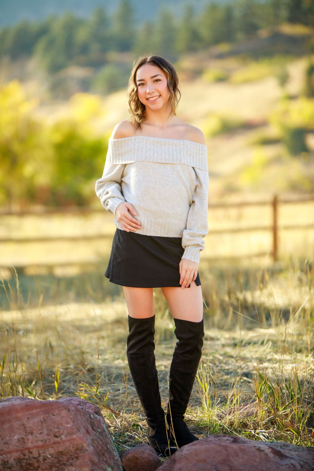 Fall fashion portrait in off-shoulder sweater and boots against mountain landscape.