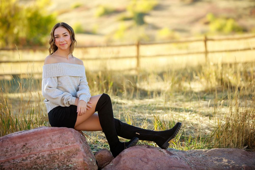 A fall portrait of someone in an off-shoulder sweater and knee-high boots sitting on a rustic rock formation.