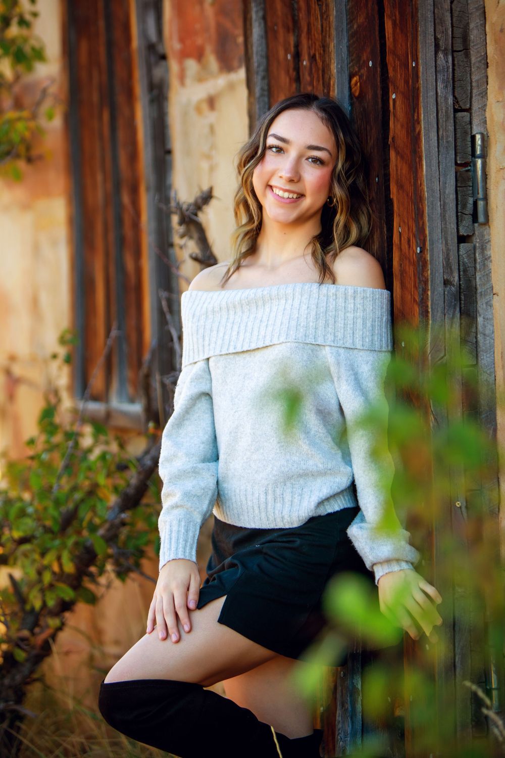A lifestyle portrait against a rustic wooden wall featuring an off-shoulder sweater and dark shorts.