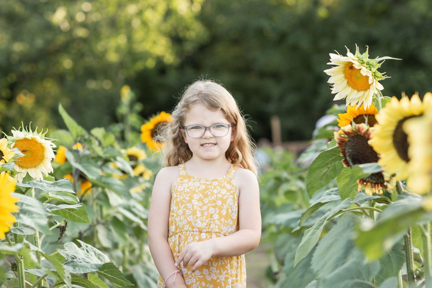 Young girl in yellow dress and glasses standing in sunflower field.