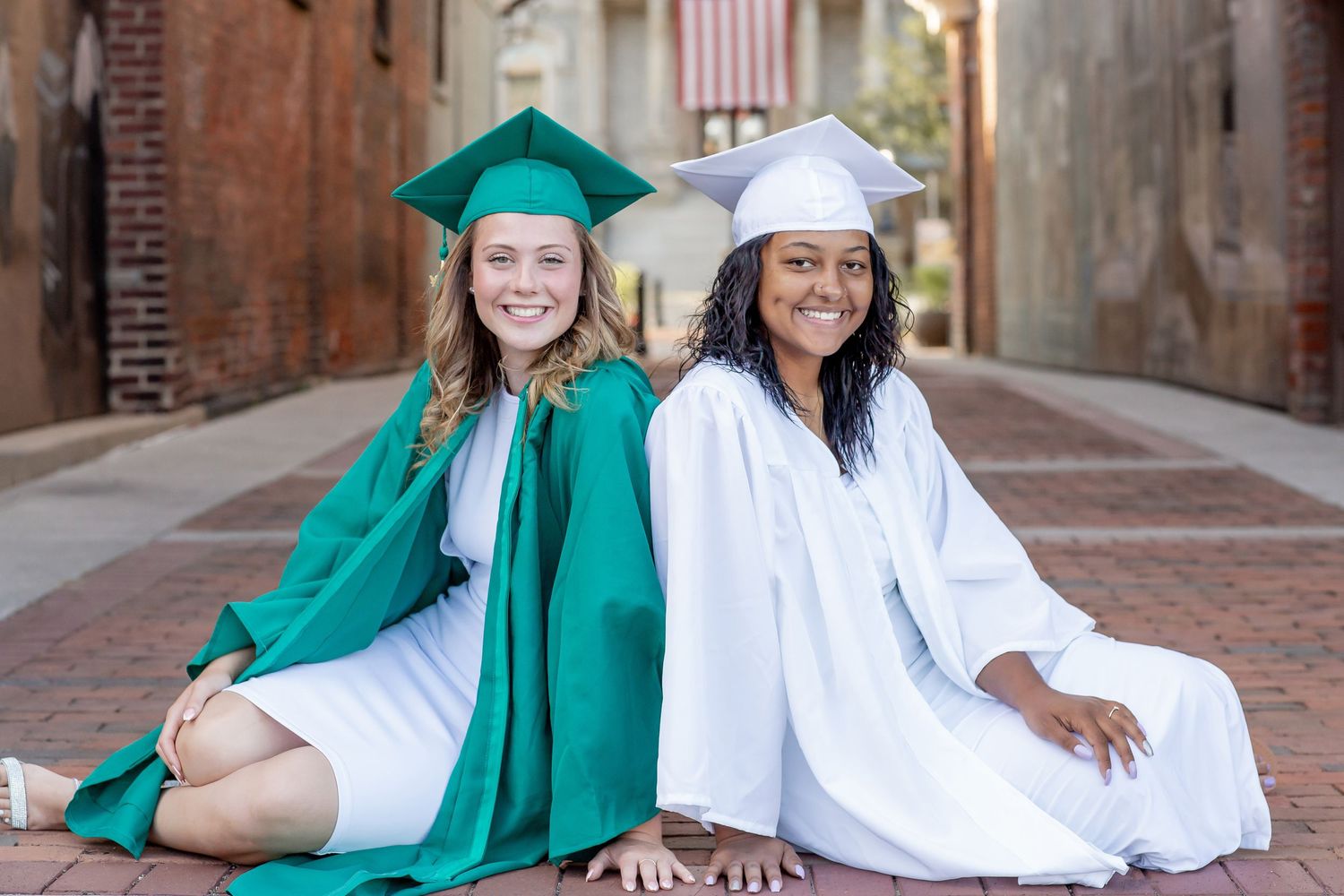 Two female graduates in green and white cap and gown sitting together.