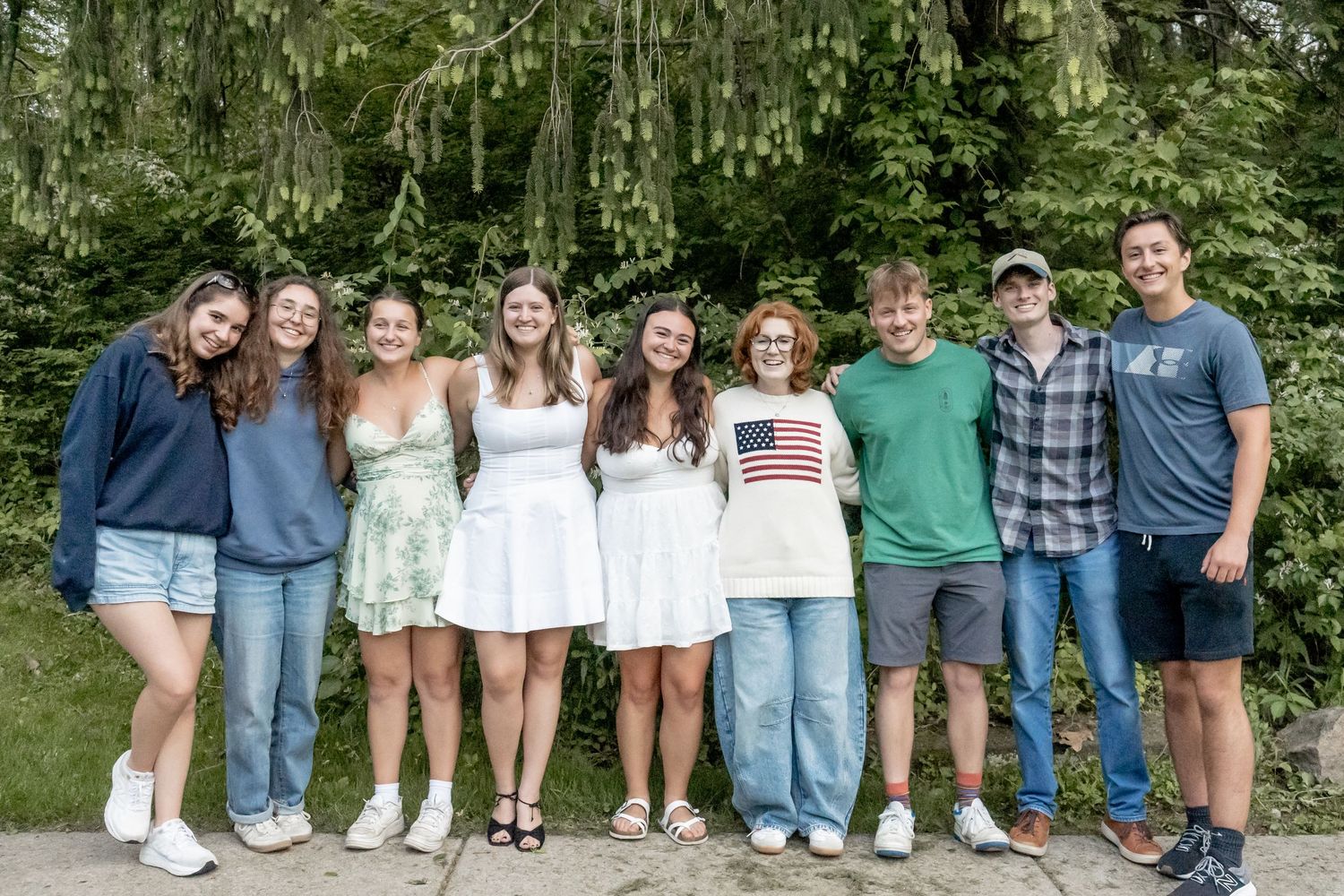 Group of nine smiling teens posing together outdoors in front of green trees on a sunny day.