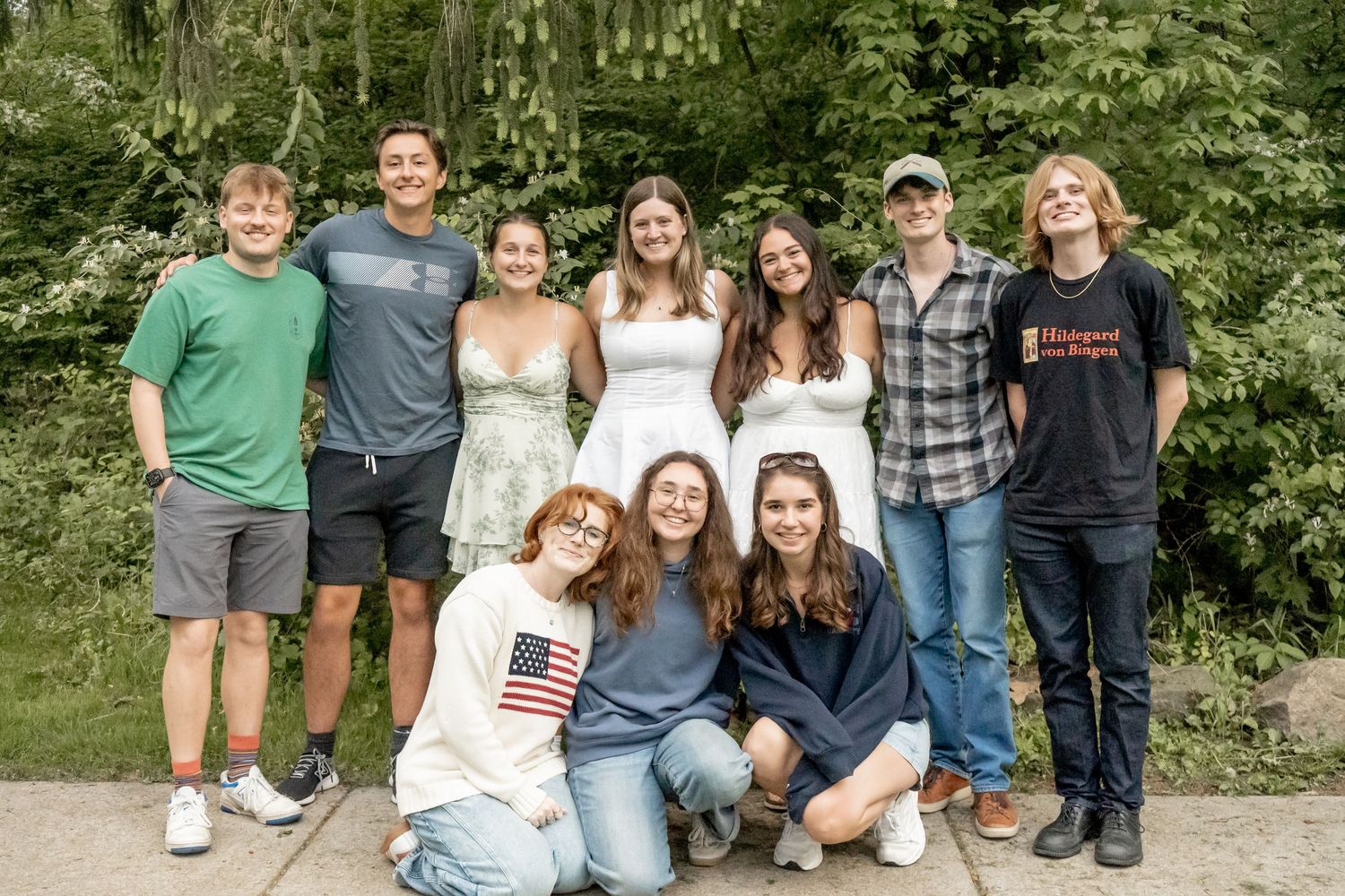 Group of ten teens posing together outdoors, some kneeling, in front of lush green trees.