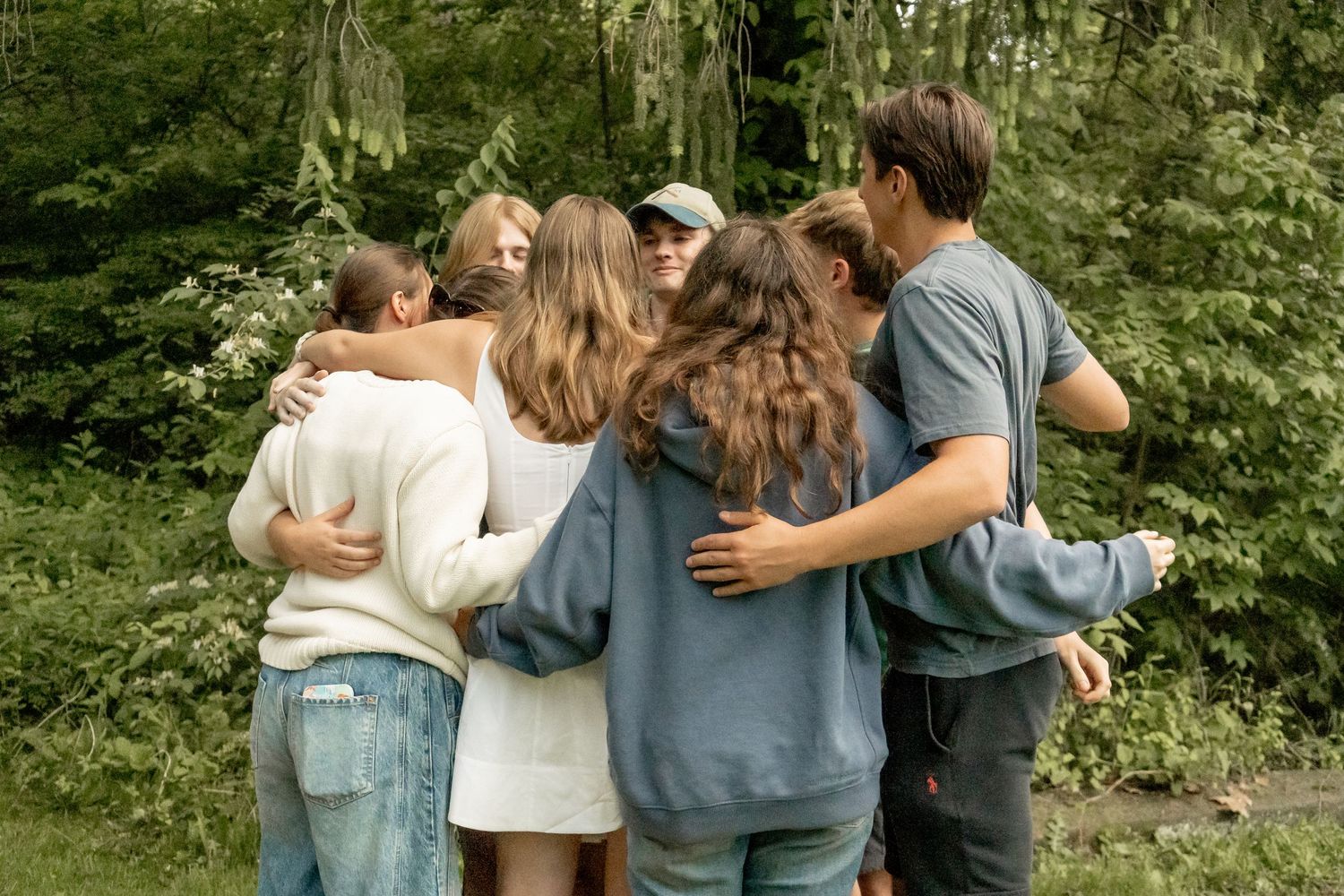 Group of teens hugging in a circle from behind surrounded by lush green trees outdoors.