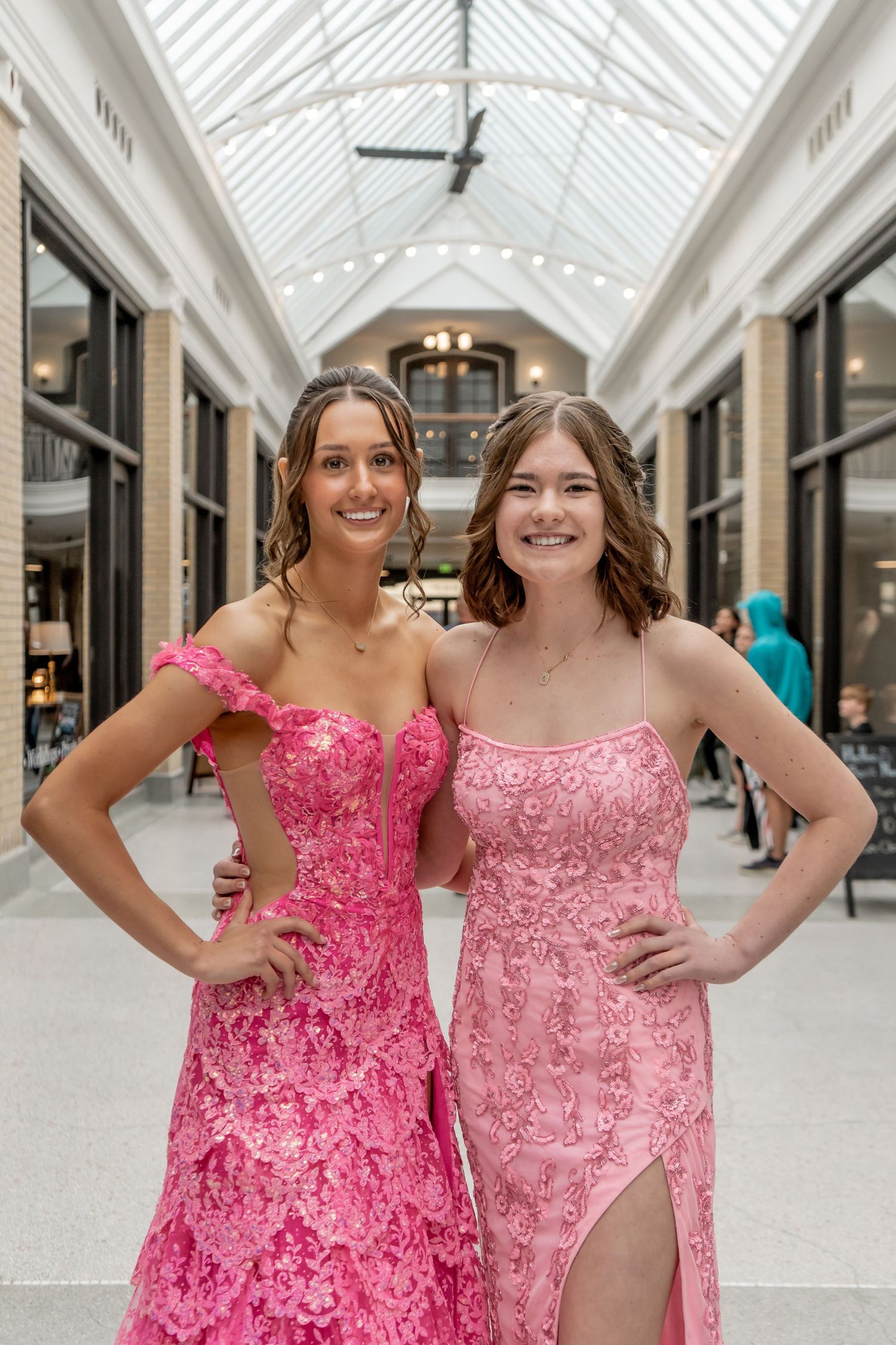 Two smiling teen girls in pink floral prom dresses posing together in an elegant indoor arcade.