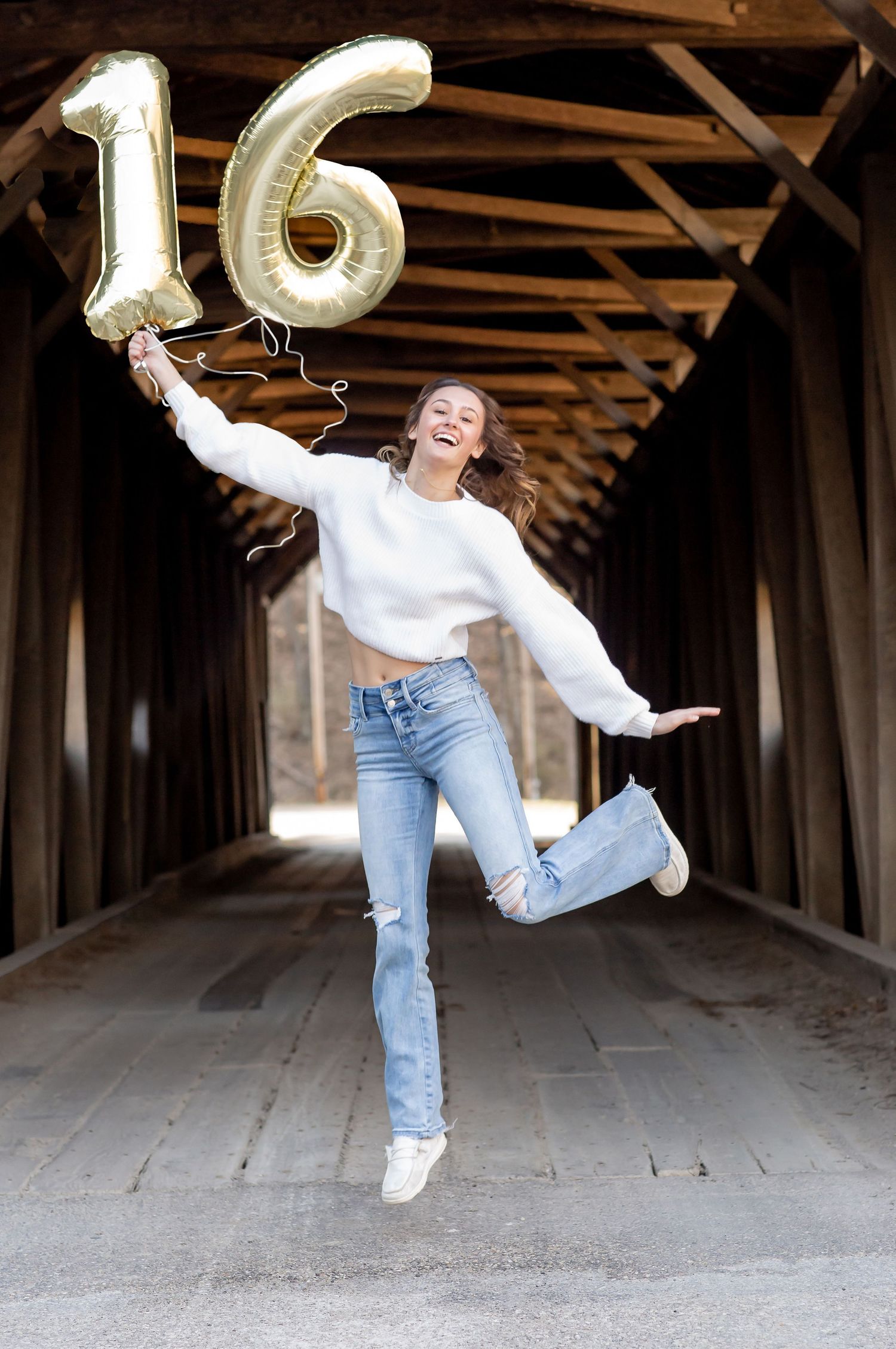 Teenage girl jumping joyfully holding gold number 16 balloons in a covered bridge.