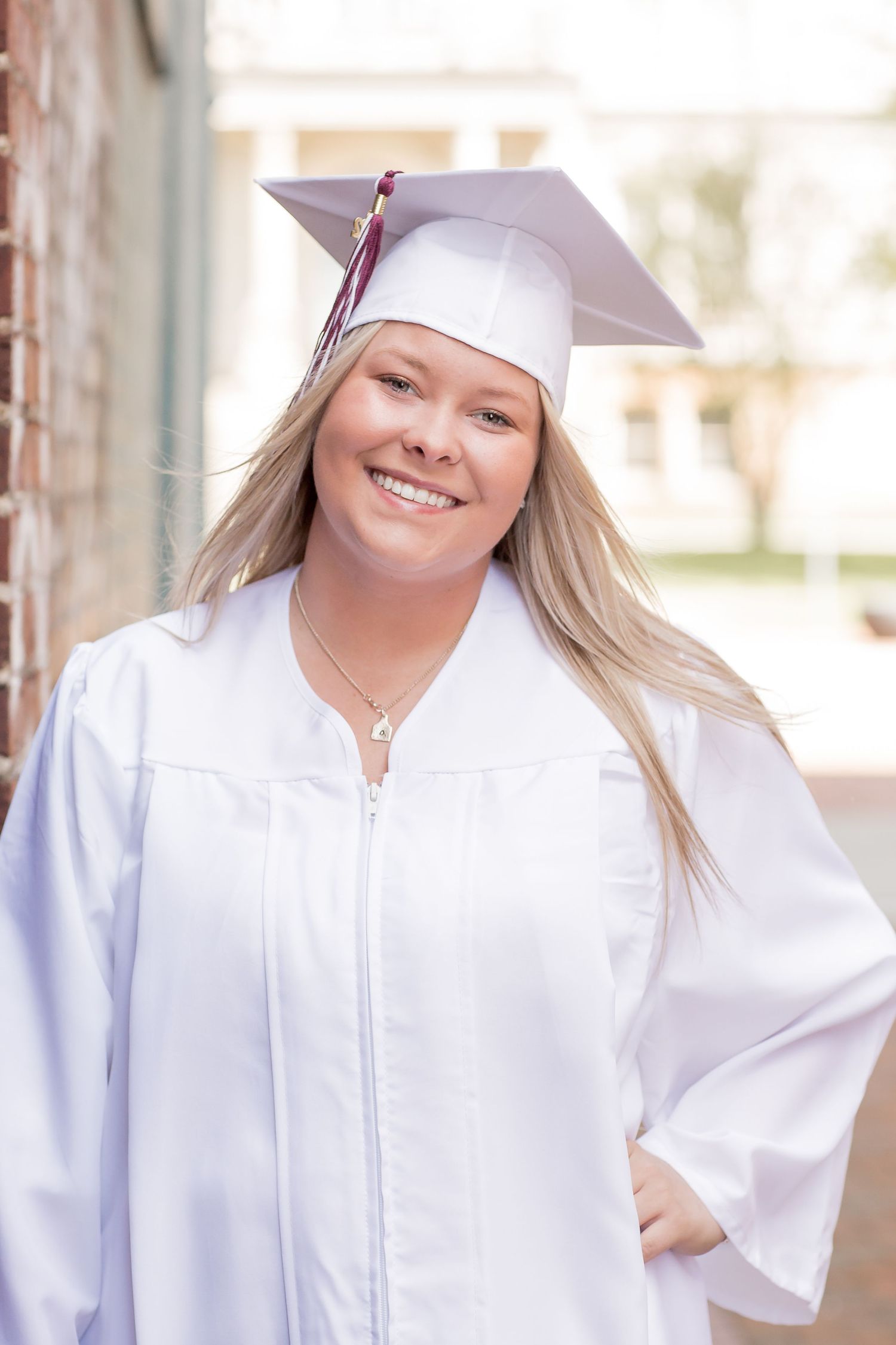 Smiling blonde graduate in white cap and gown posing outdoors.