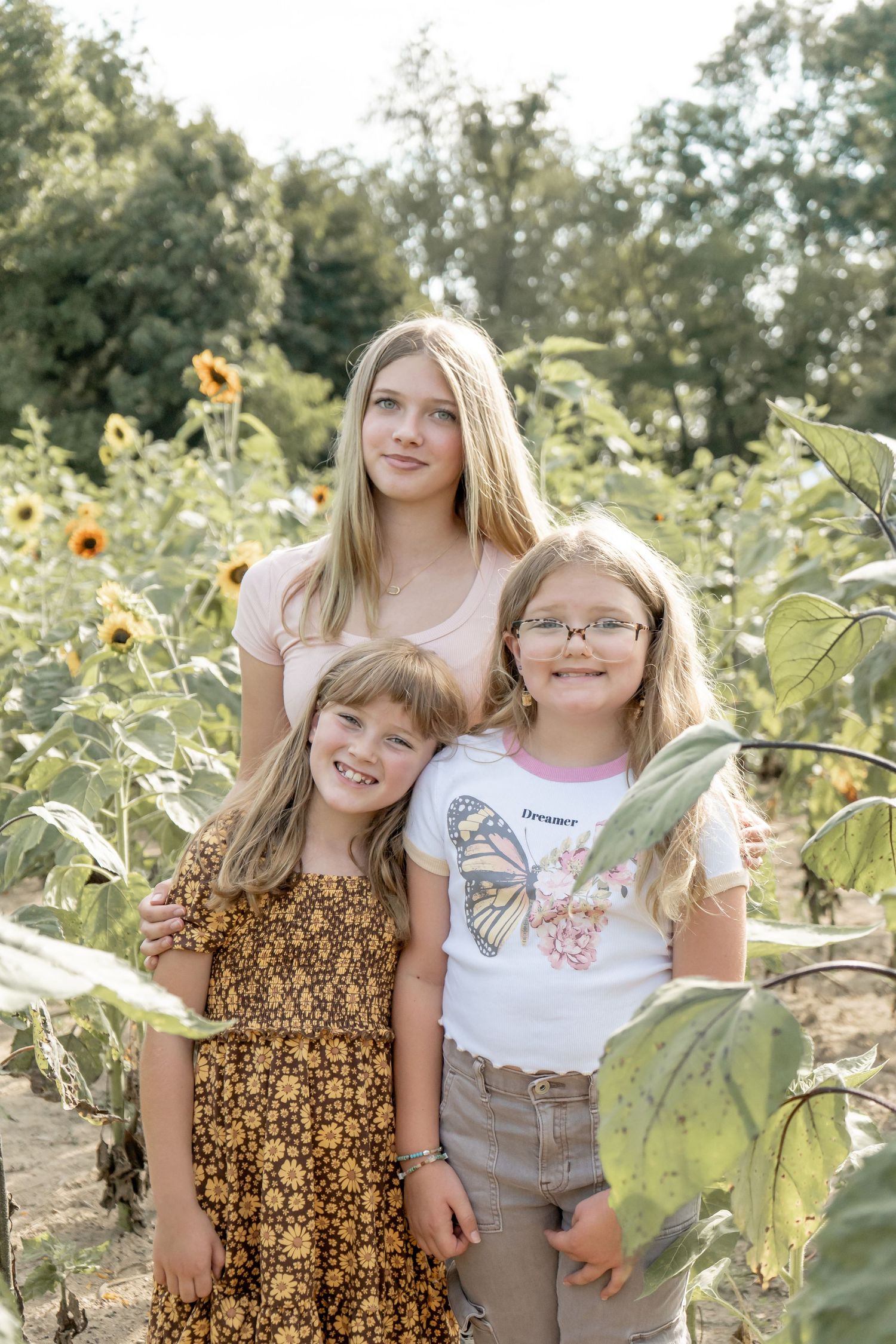 Three sisters smiling together in a sunflower field on a sunny day.